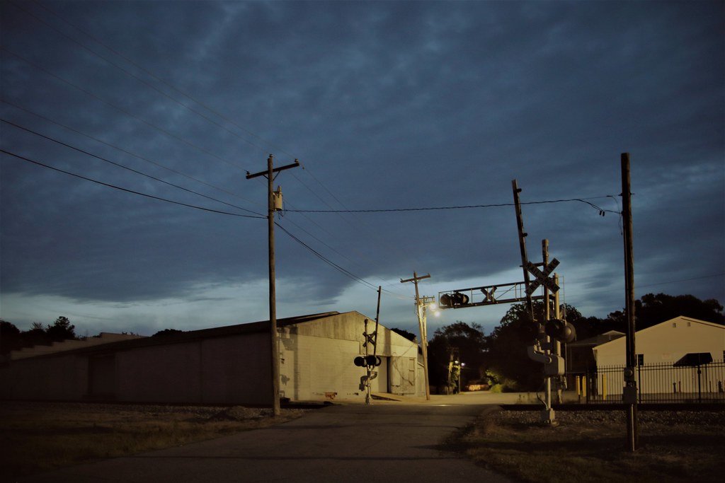 Nighttime view of a railroad crossing with warning signals, utility poles, and industrial buildings under a cloudy sky.