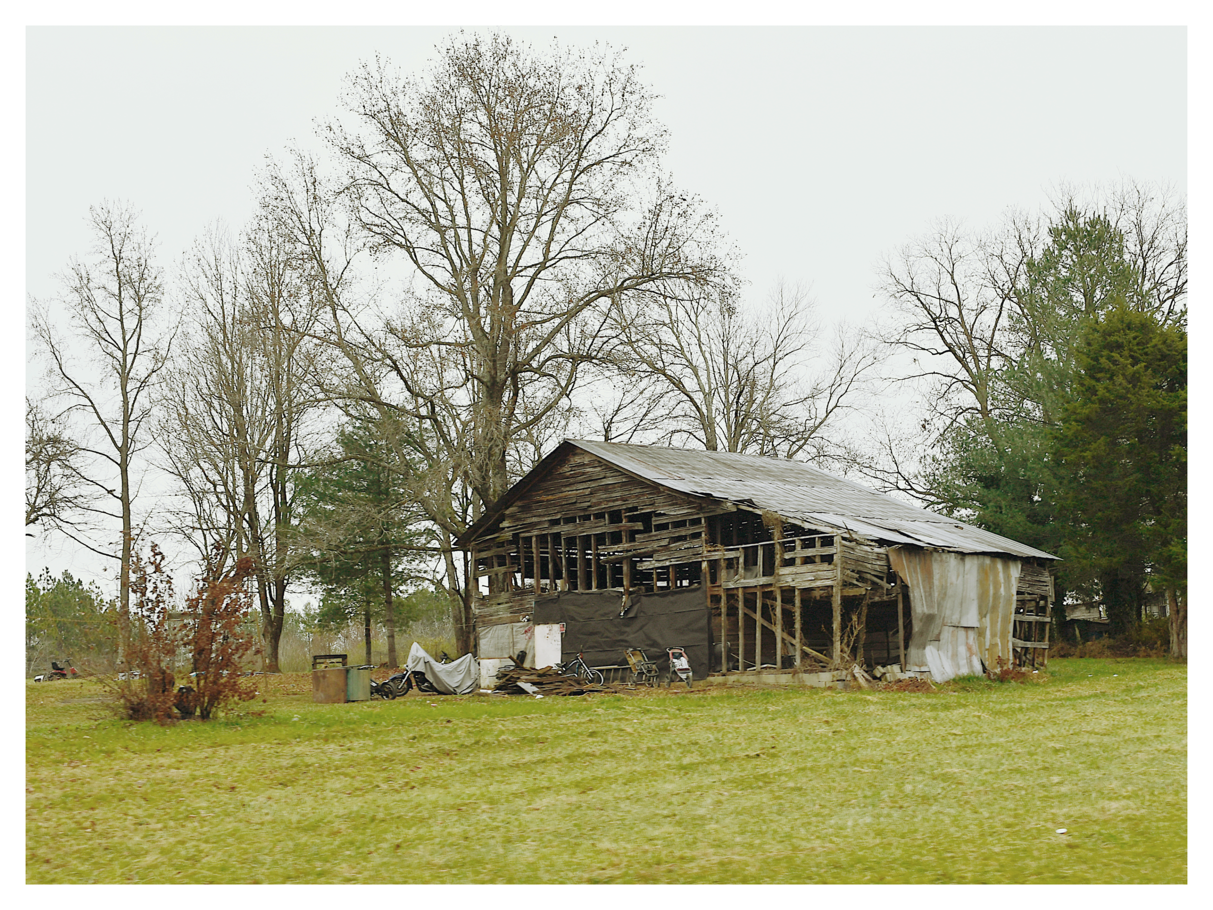 An old, dilapidated wooden barn with a tin roof, surrounded by leafless trees, with some motorcycles and debris in front of it, on a grassy area under an overcast sky.