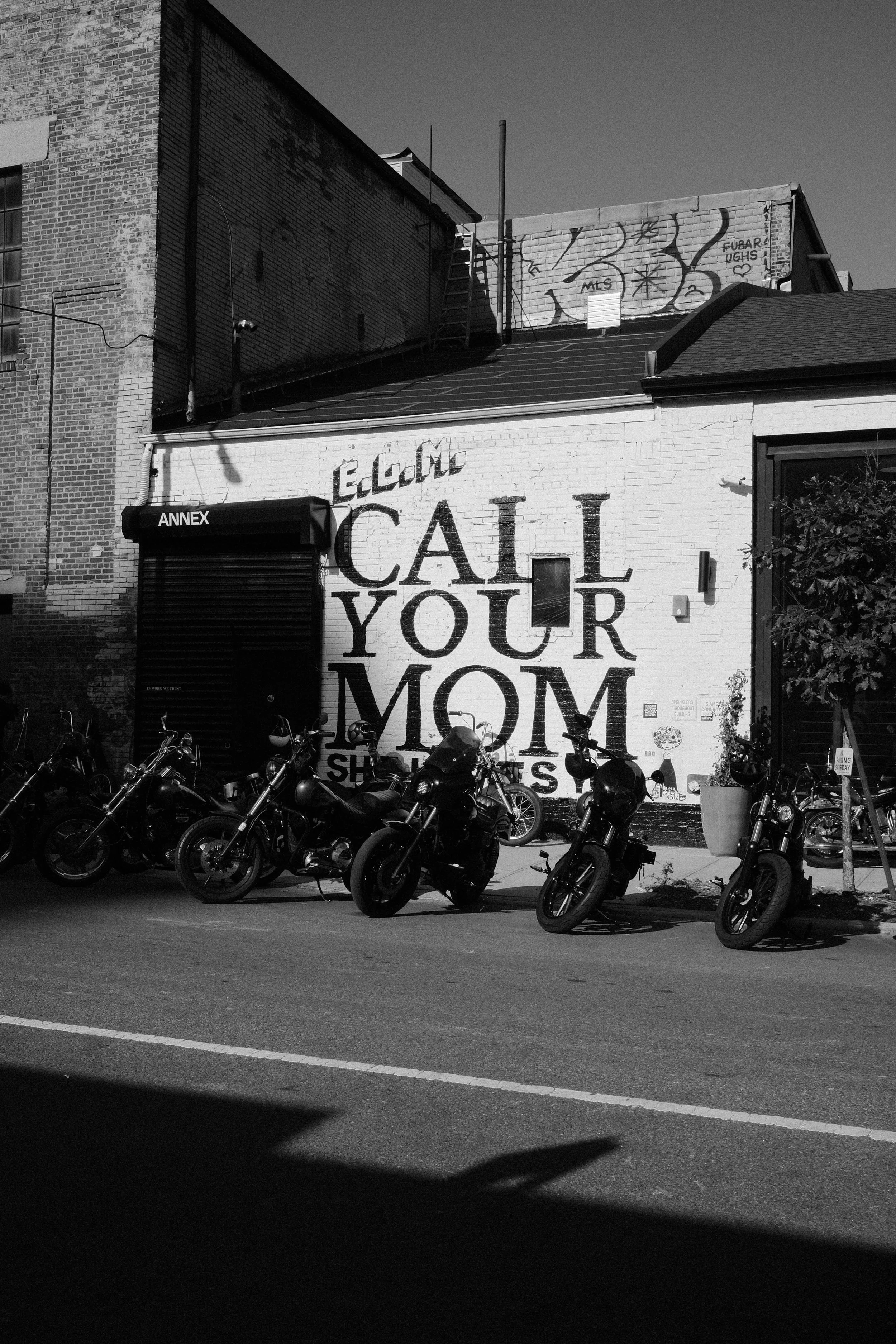 Black and white photo of a building with a large mural that says "Call Your Mom" and a row of parked motorcycles in front.