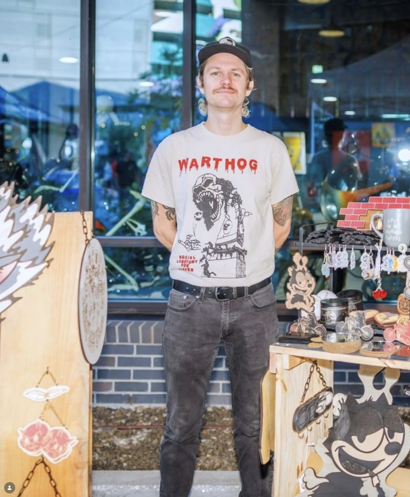 A young man with a mustache, wearing a black cap and a white T-shirt with 'WARTHOG' written on it, stands behind a display table at an indoor market. The table has various small crafts, including stickers and keychains, and large cartoon character cutouts. The background shows a glass window with reflections of other market stalls and people.