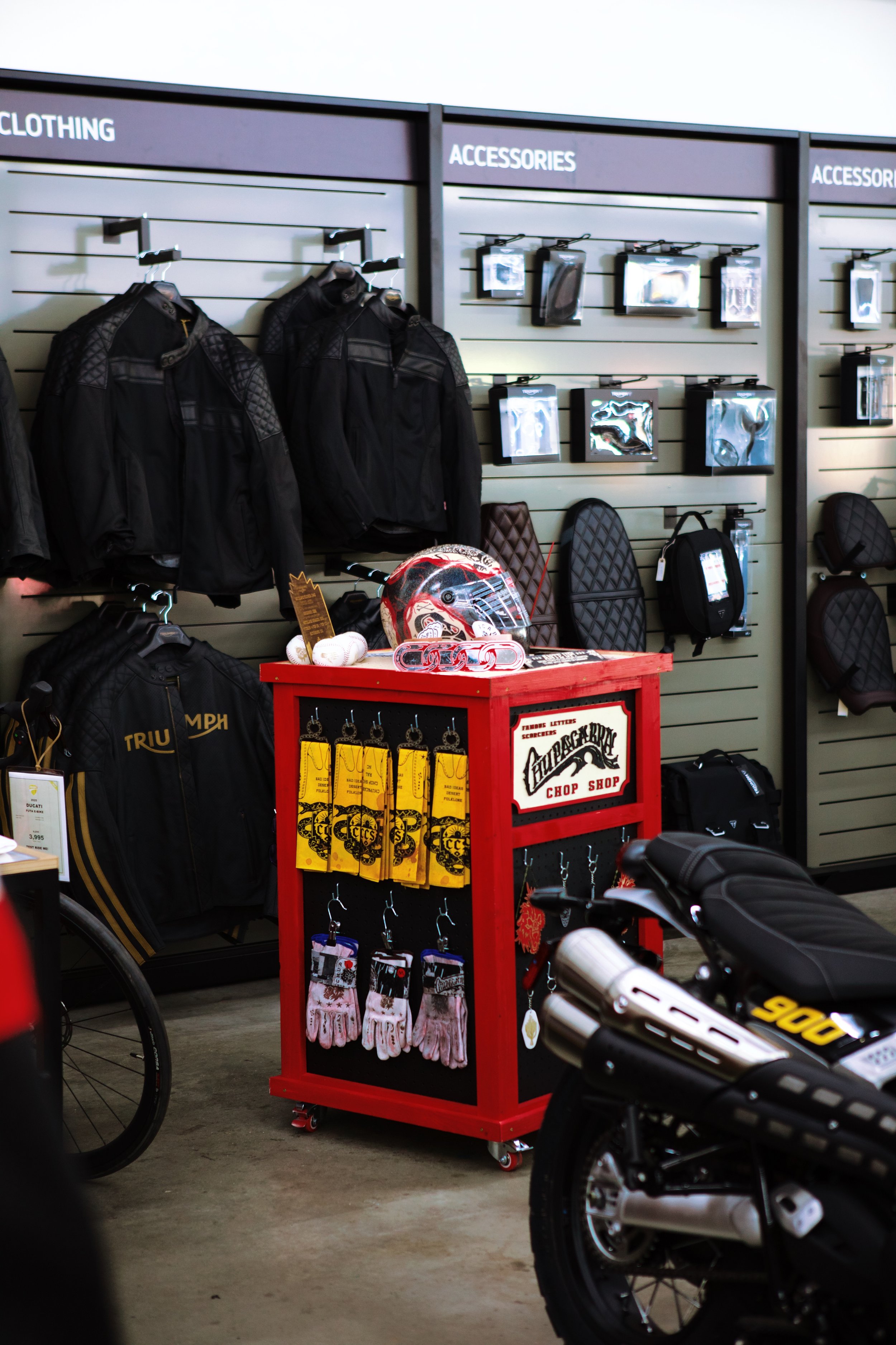 A retail store display showcasing motorcycle jackets, accessories, gloves, and other gear, with a motorcycle in the foreground.