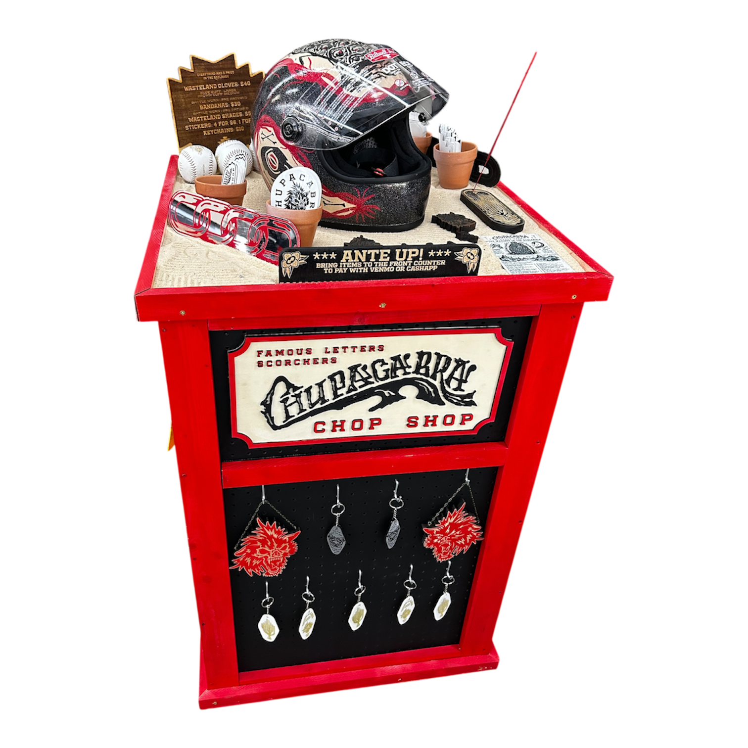 A red display stand with various items including a black helmet, toy baseballs, keychains, and a sign, at a shop called Fur Sacrifice Chop Shop which sells scarceries and accessories.