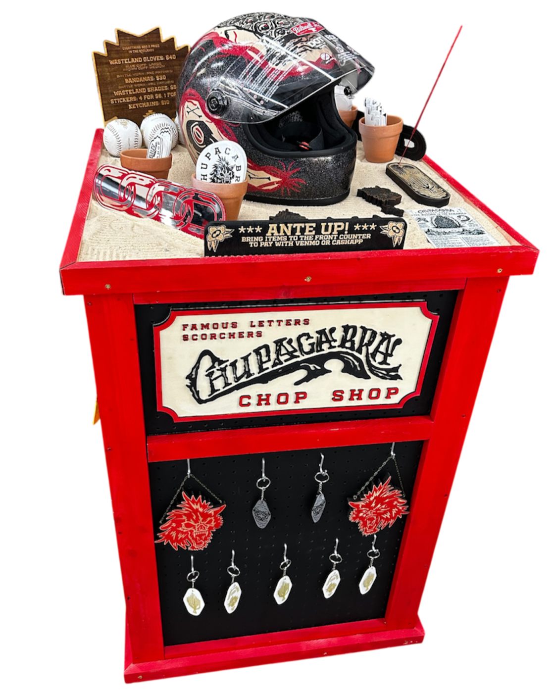 Red display stand for a fictional chop shop called Chupacabra Chop Shop, decorated with a Chupacabra head decorations and hanging products, with various accessories and a helmet on top.