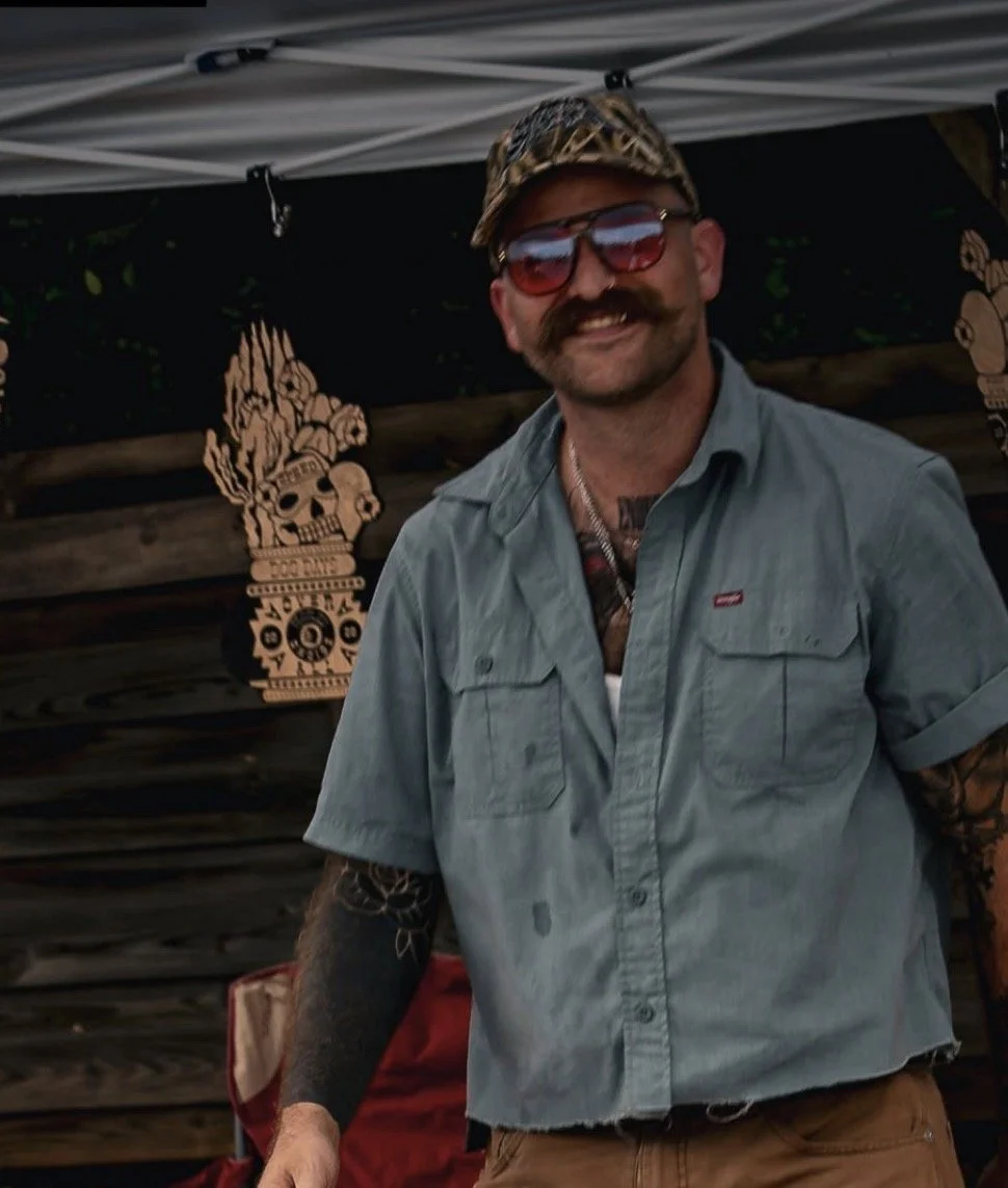 A man with a beard and mustache wearing sunglasses, a camouflage cap, a light blue short-sleeve button-up shirt, and a tattoo on his right forearm. He is smiling and standing in front of a wooden background with Halloween-themed decorations, including a large wooden skull with flames and other designs.