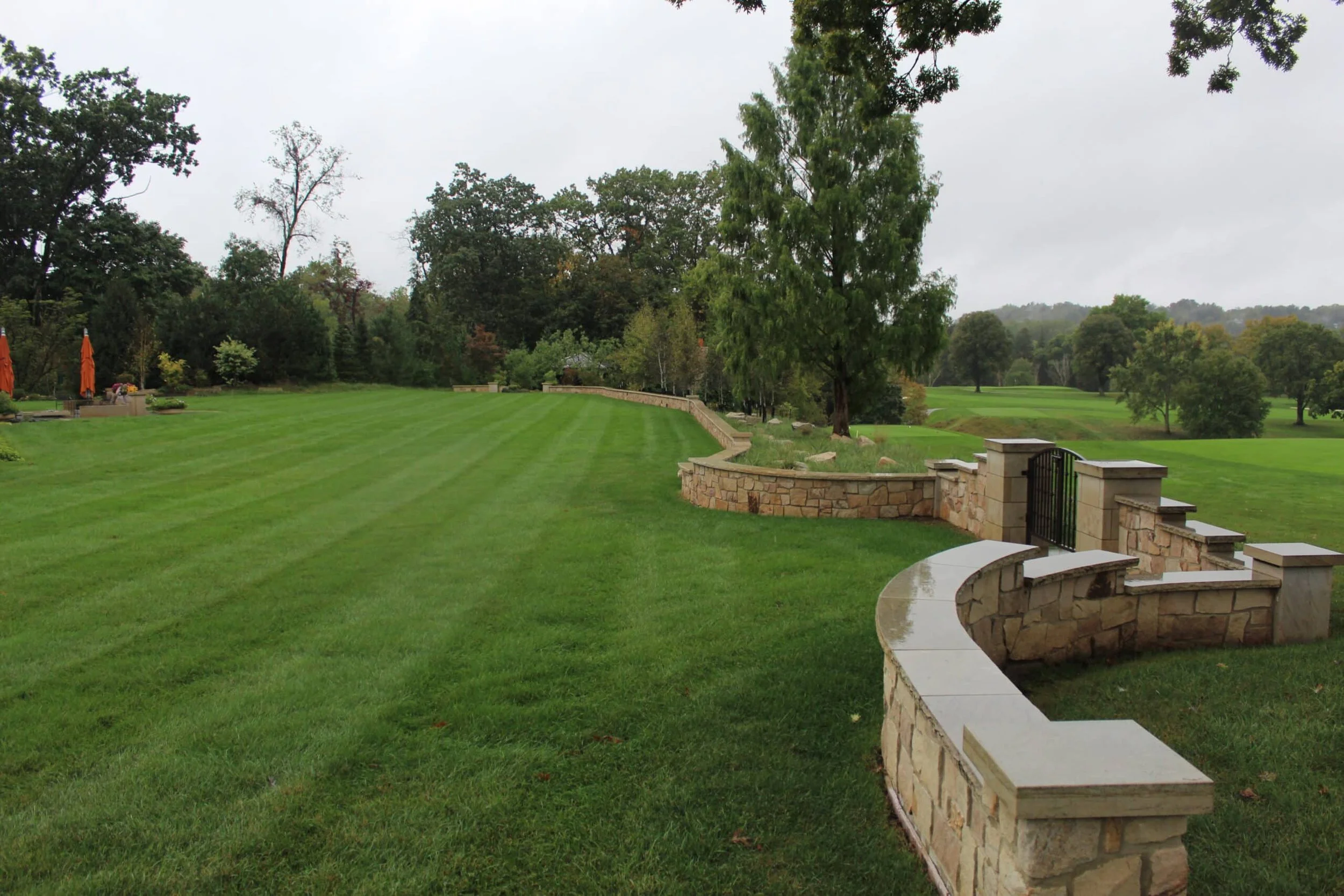 A large, green, well-manicured lawn with a curved stone wall and black metal gate in the foreground. Tall trees and cloudy sky in the background.