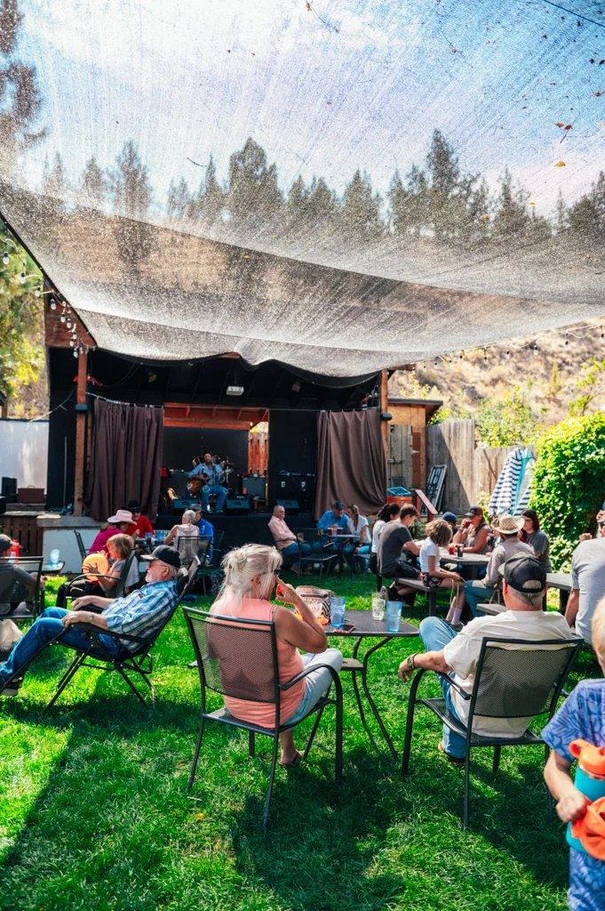 People sitting on outdoor chairs and tables in a backyard, watching a live music performance on a small stage with a band and a guitarist, under a large sunshade, with trees and wooden fences in the background.