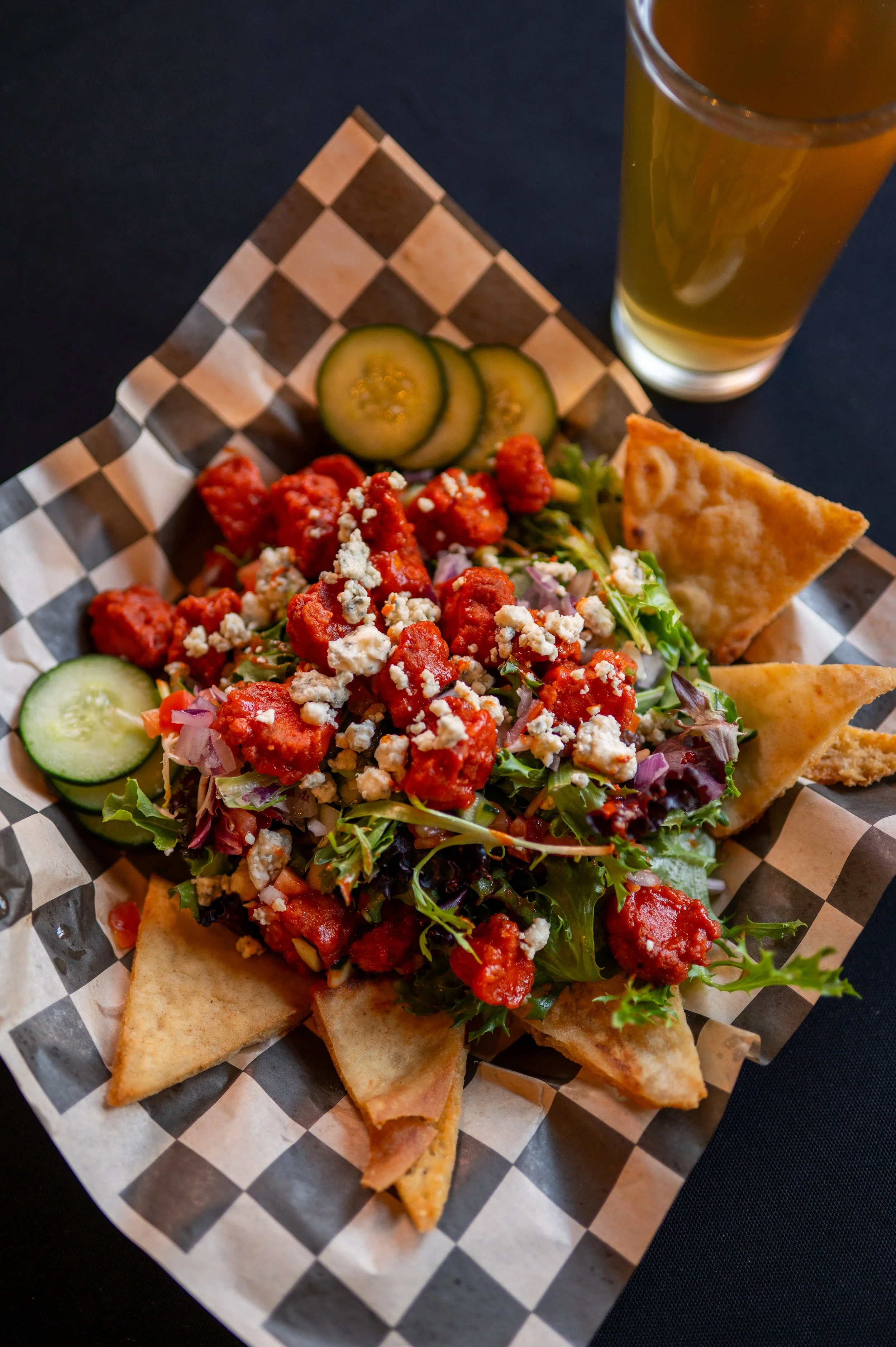A serving of nachos on a checkered paper-lined tray includes tortilla chips topped with chopped lettuce, diced onions, crumbled blue cheese, and spicy meat sauce, with slices of cucumber and a glass of beer in the background.