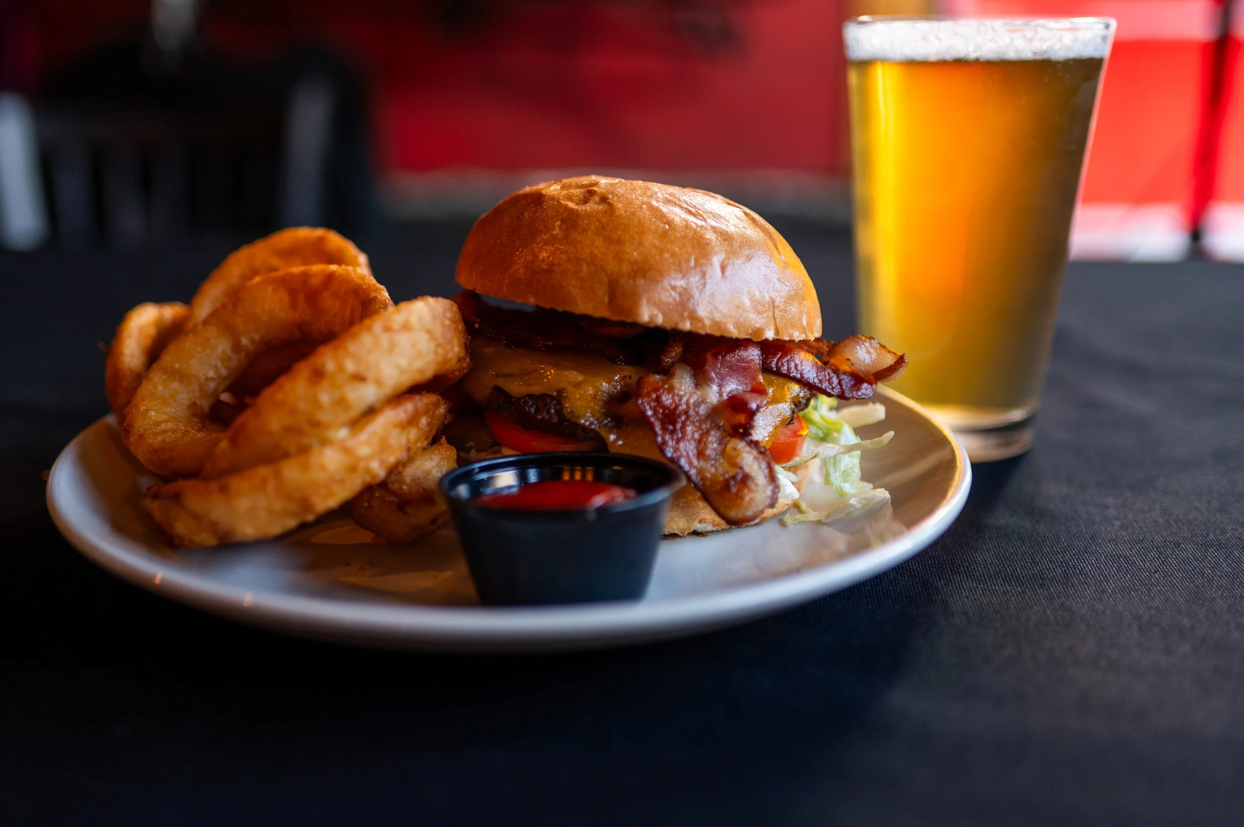 A plate of a bacon cheeseburger with onion rings and a small cup of dipping sauce, alongside a glass of beer on a dark table.