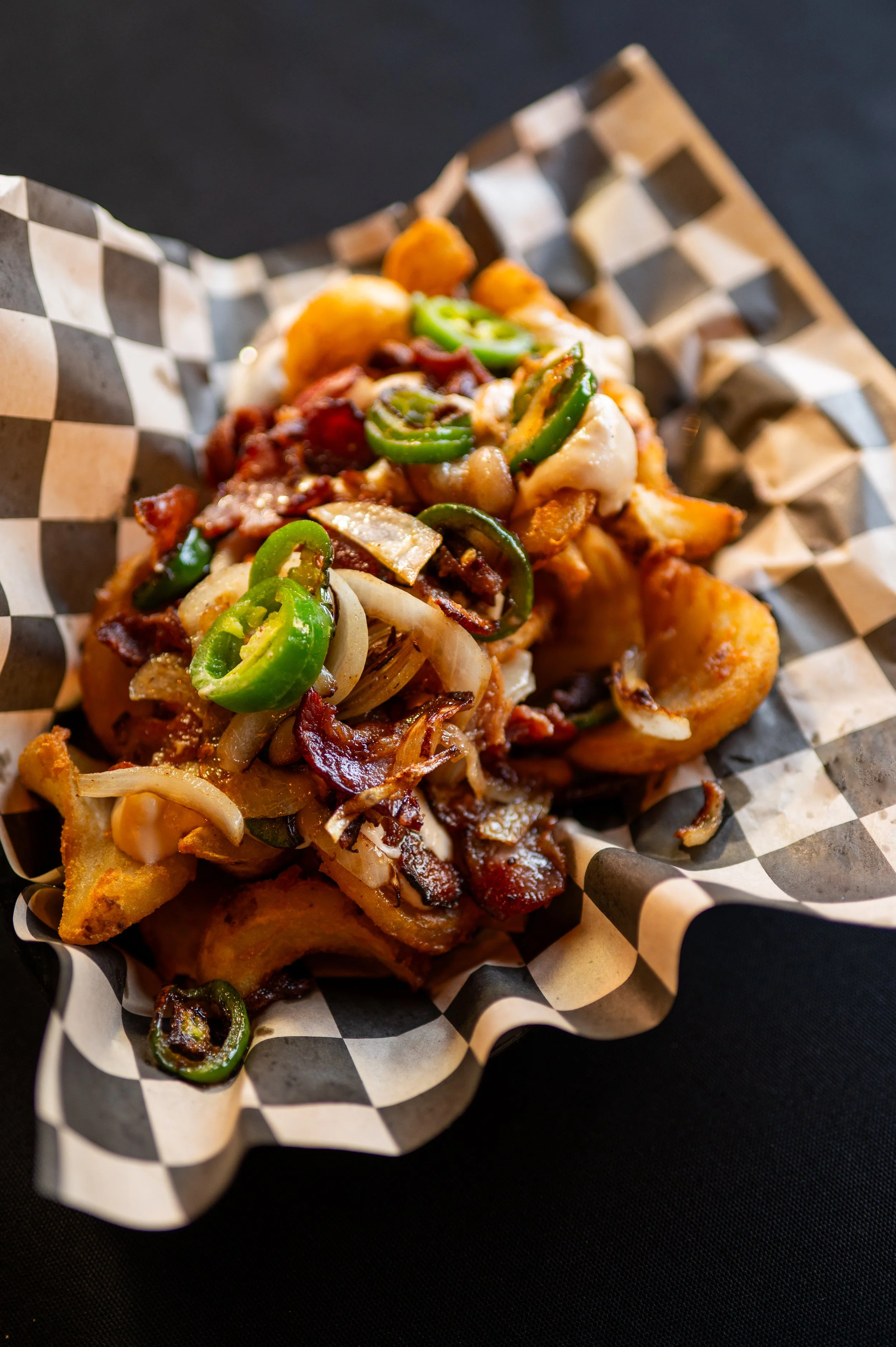 Close-up of a slice of loaded pizza with green peppers, onions, bacon, and melted cheese, served on checkered paper.