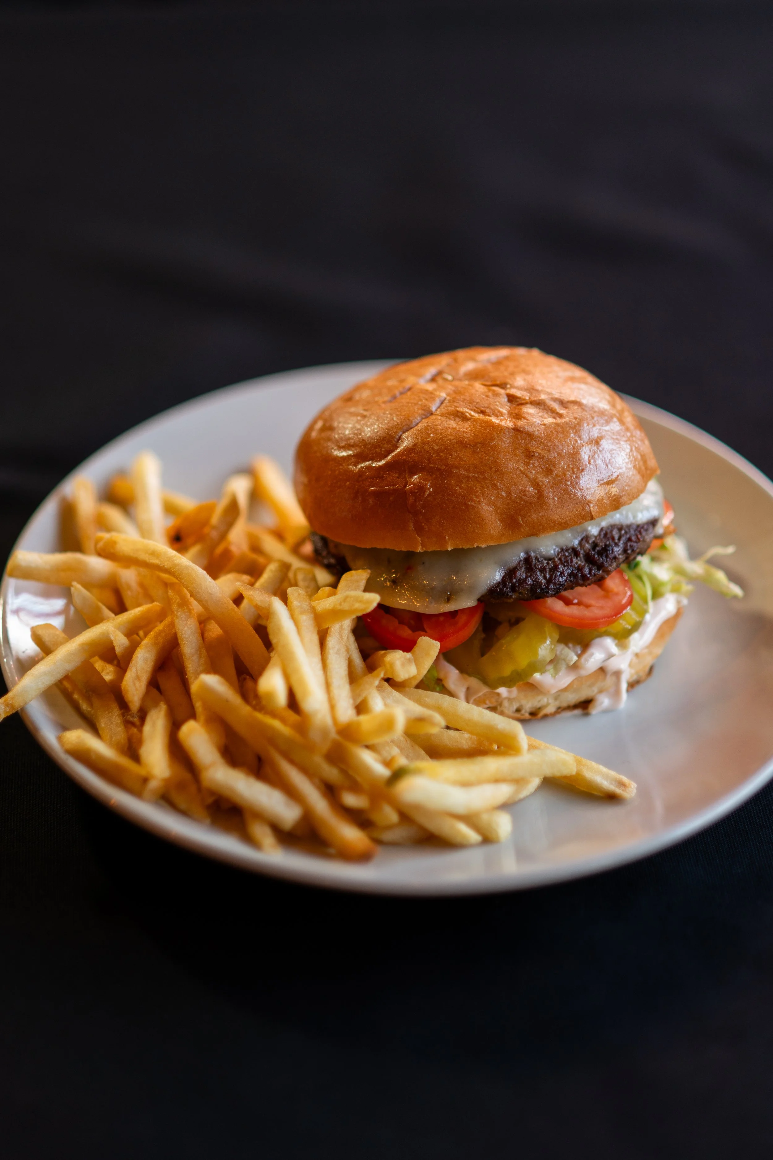 Close-up of a cheeseburger with lettuce, tomato, pickles, and melted cheese on a sandwich bun, served with a side of French fries on a white plate against a dark background.