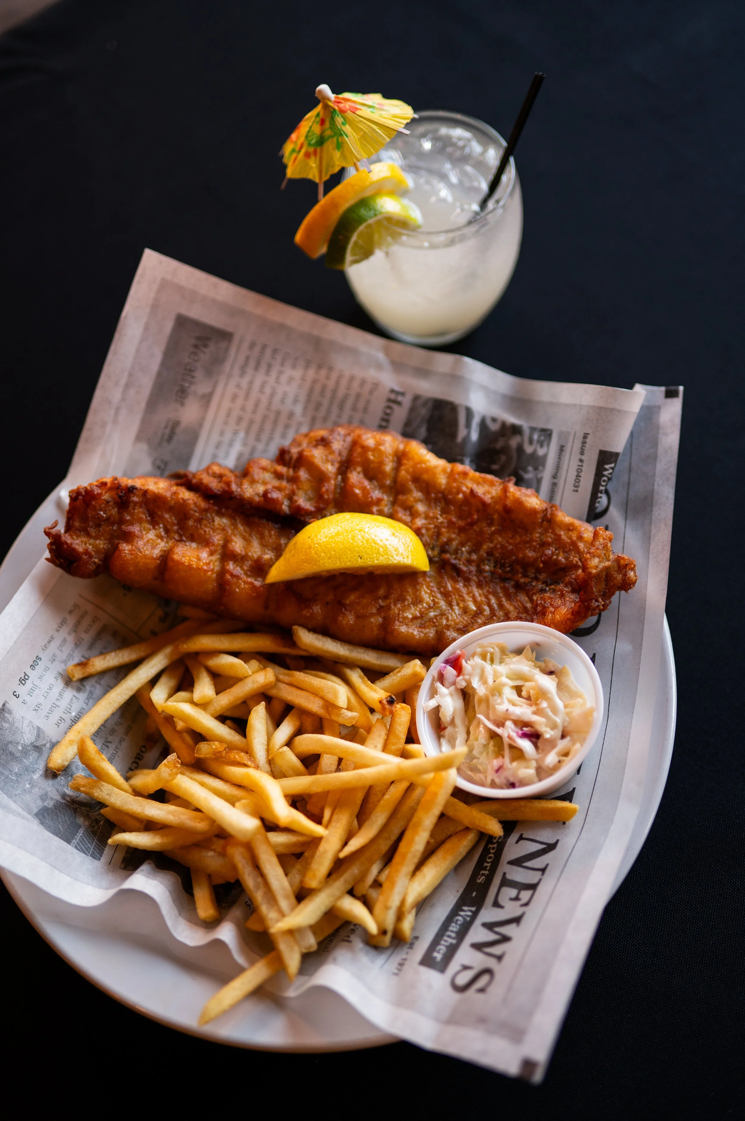 Fried fish fillet with a lemon wedge, French fries, coleslaw, and a side of creamy dipping sauce on a paper-lined tray. In the background, a glass of lemonade with lemon slices and a small yellow parasol.