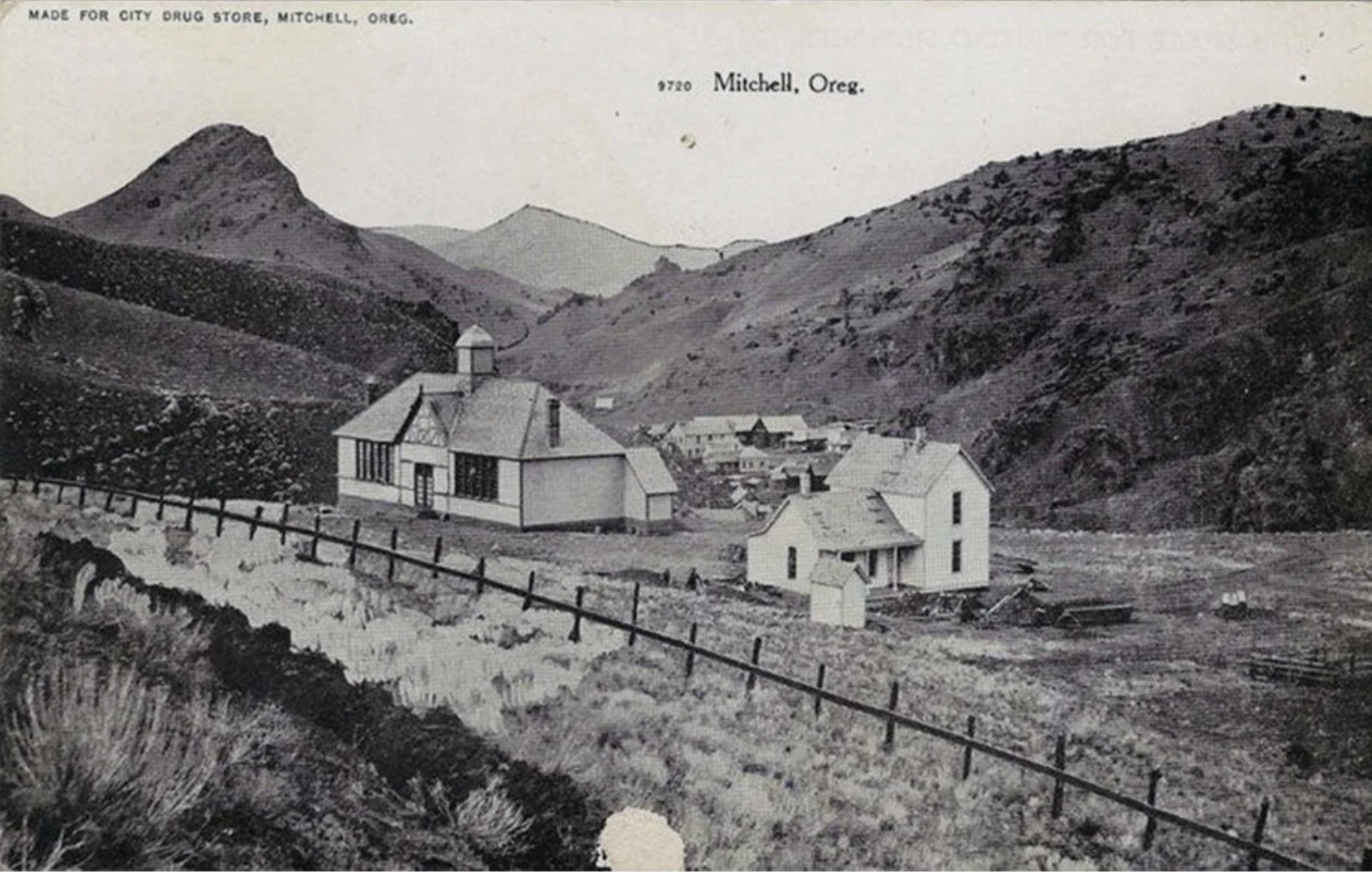 Black and white photo of a rural landscape with mountains in the background, a few houses, and a fence along a small stream. The photo is labeled 'Made for City Drug Store, Mitchell, Oreg.'