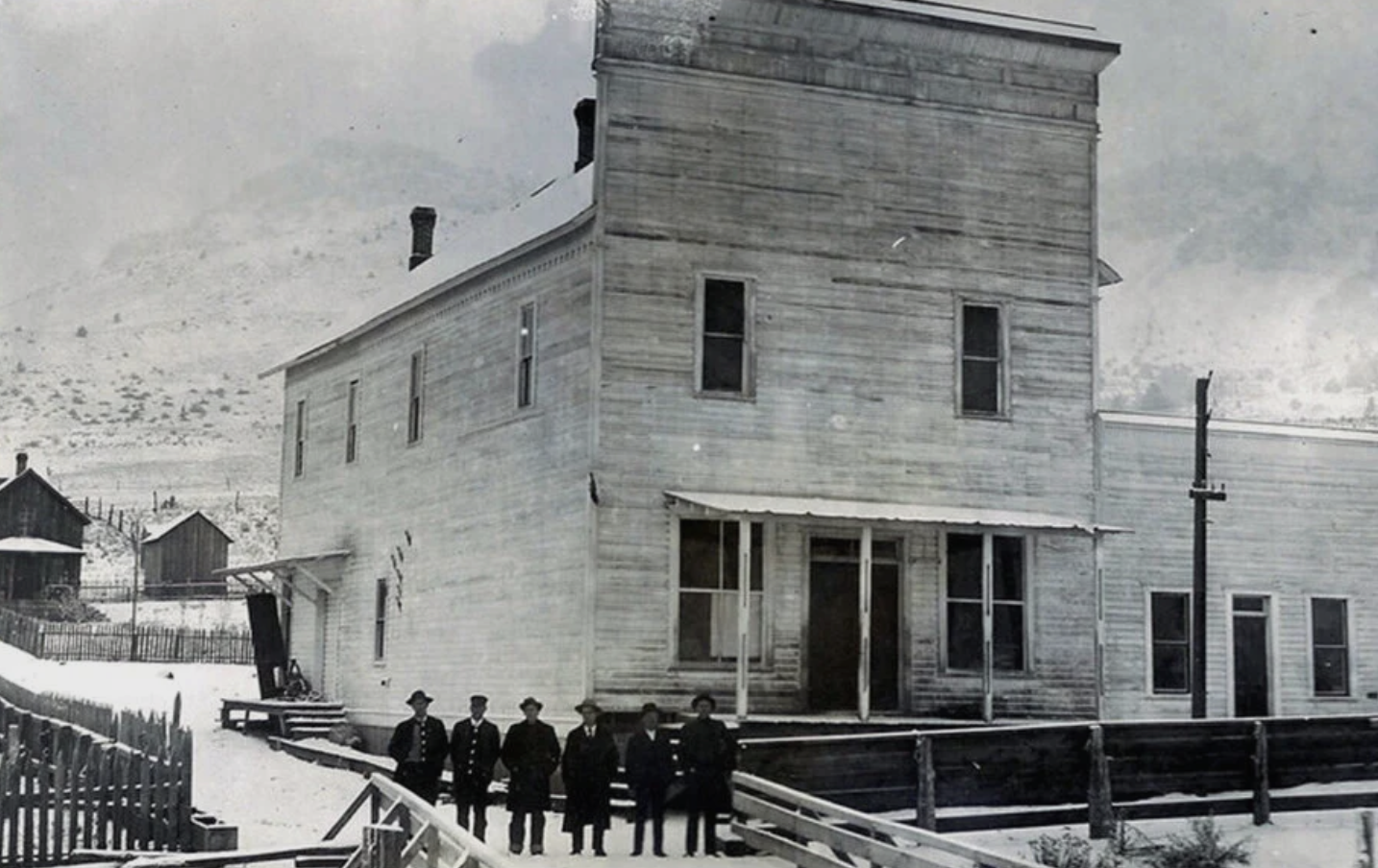 Old black and white photo of a large wooden house with five men standing in front of it. The house is weathered, with peeling paint, and is set in a snowy landscape with mountains in the background.