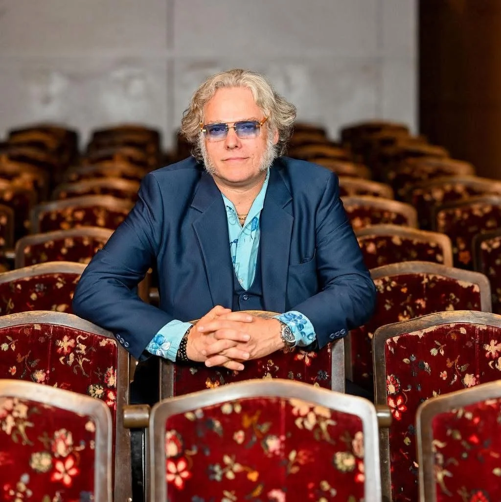 A man with curly blonde hair, blue sunglasses, and a blue suit sitting at a table in an empty auditorium with red floral-patterned chairs.