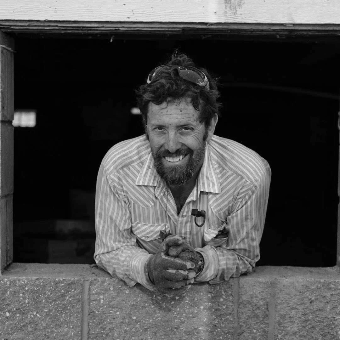 A man with a beard and curly hair smiling, wearing sunglasses on his head and a striped shirt, leaning out of a small opening in a wall, holding a small bird in his gloved hands.