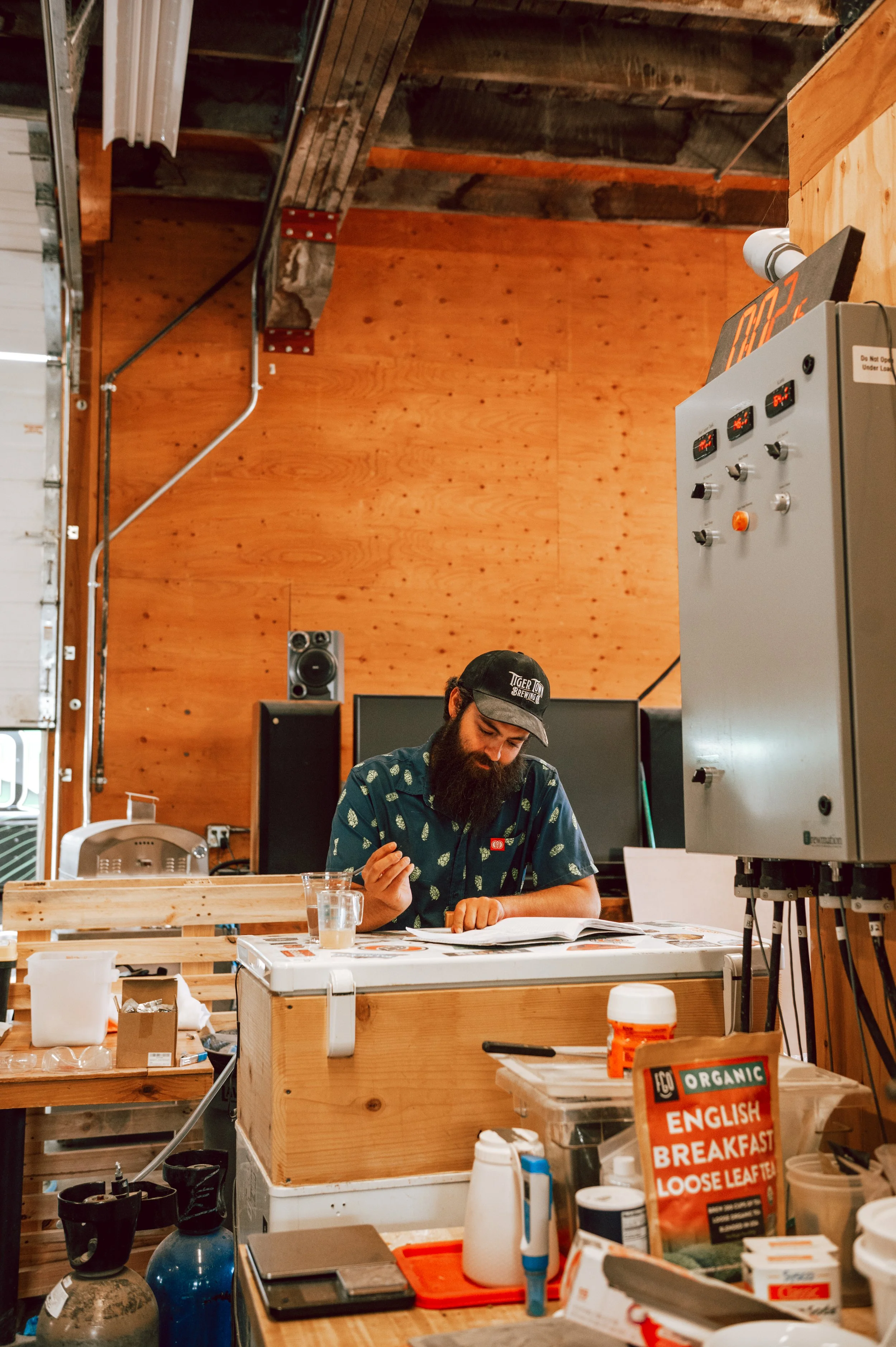 Man with a beard wearing a baseball cap and patterned shirt sitting at a cluttered wooden table in a workshop or studio, reading a book or magazine, with art supplies and containers on the table, and wooden walls and ceiling in the background.