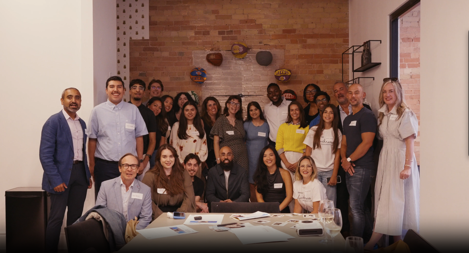 Group photo of diverse people at a social or professional gathering in a room with a brick and white wall background, some standing and some sitting, with tables, papers, and glasses visible in the foreground.