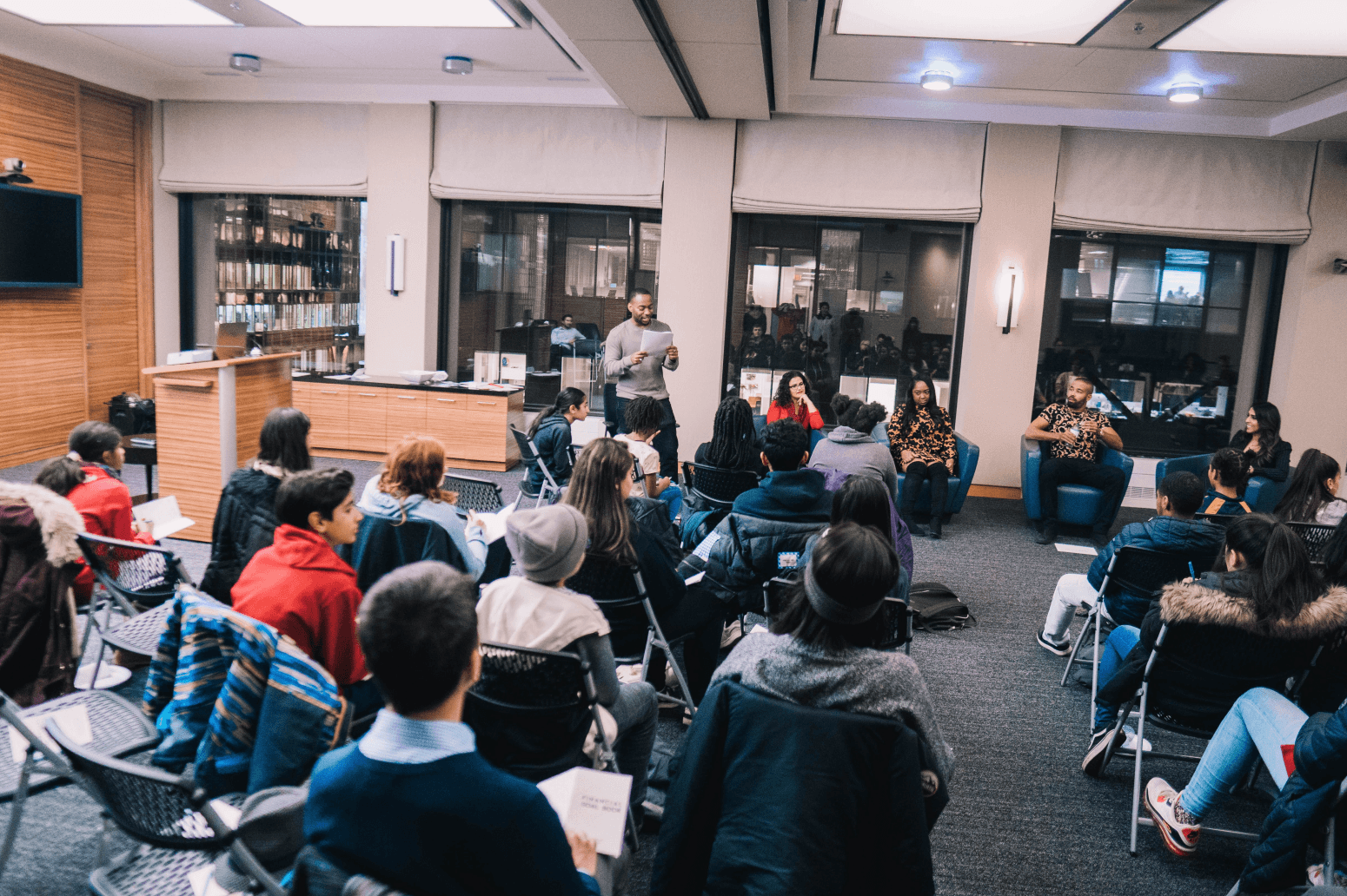 People attending a panel discussion in a conference room, with a speaker reading from notes, seated in front of an audience that is listening attentively.
