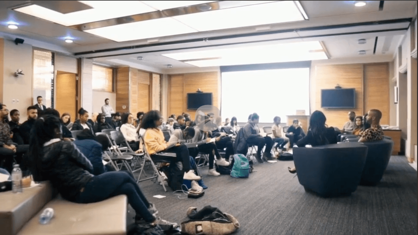 A large group of people attending a conference or seminar in a room with wooden panel walls, some seated on chairs and a few on couches, listening to a speaker at the front.
