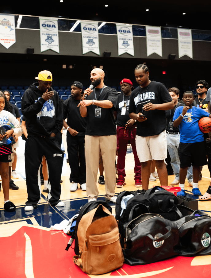 Coaching staff and young players at a basketball court during an event or team gathering, with backpacks on the floor and banners hanging from the ceiling.