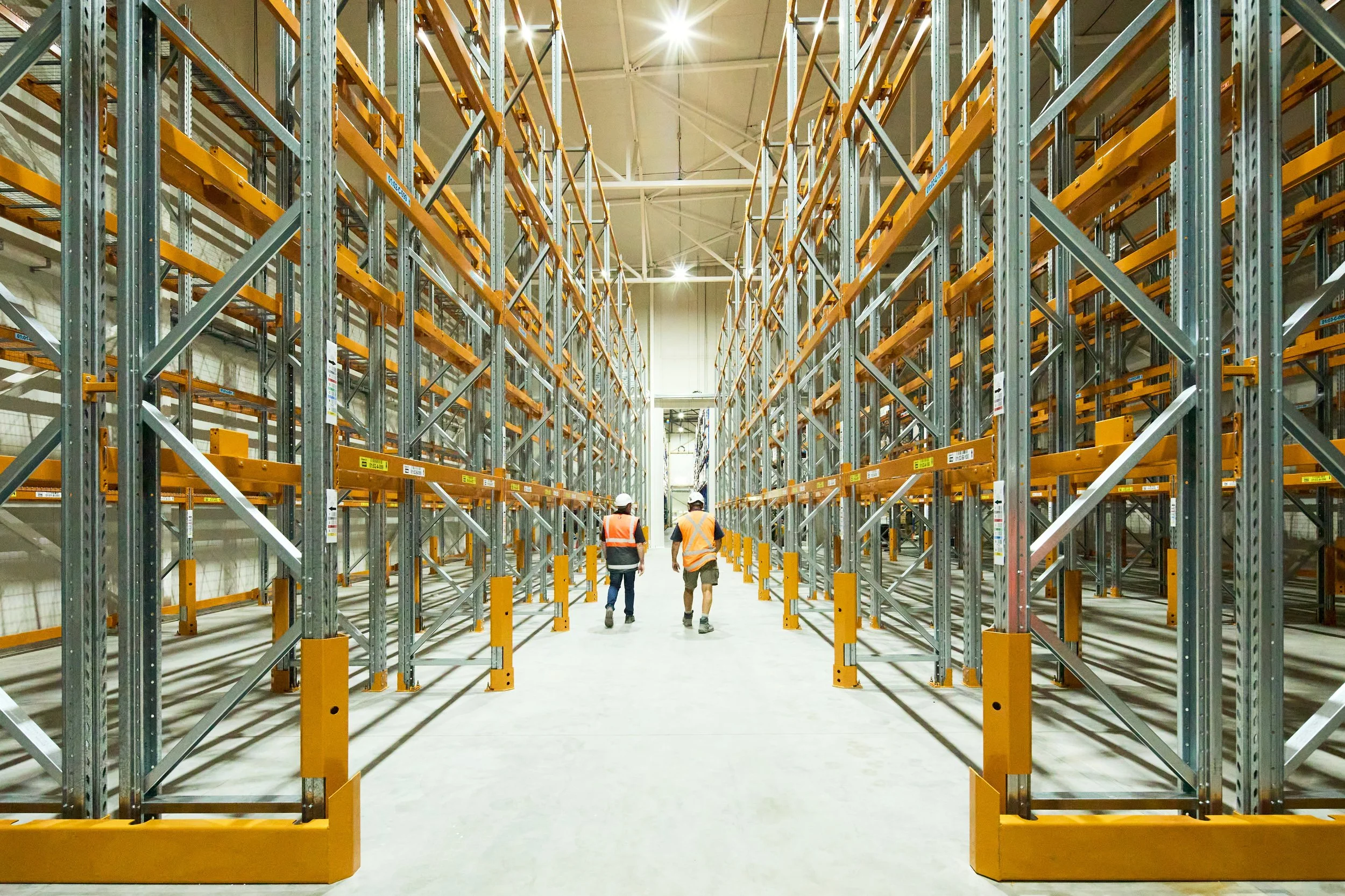 Three workers wearing safety vests and helmets walking down an aisle in a large warehouse with empty metal shelving units.