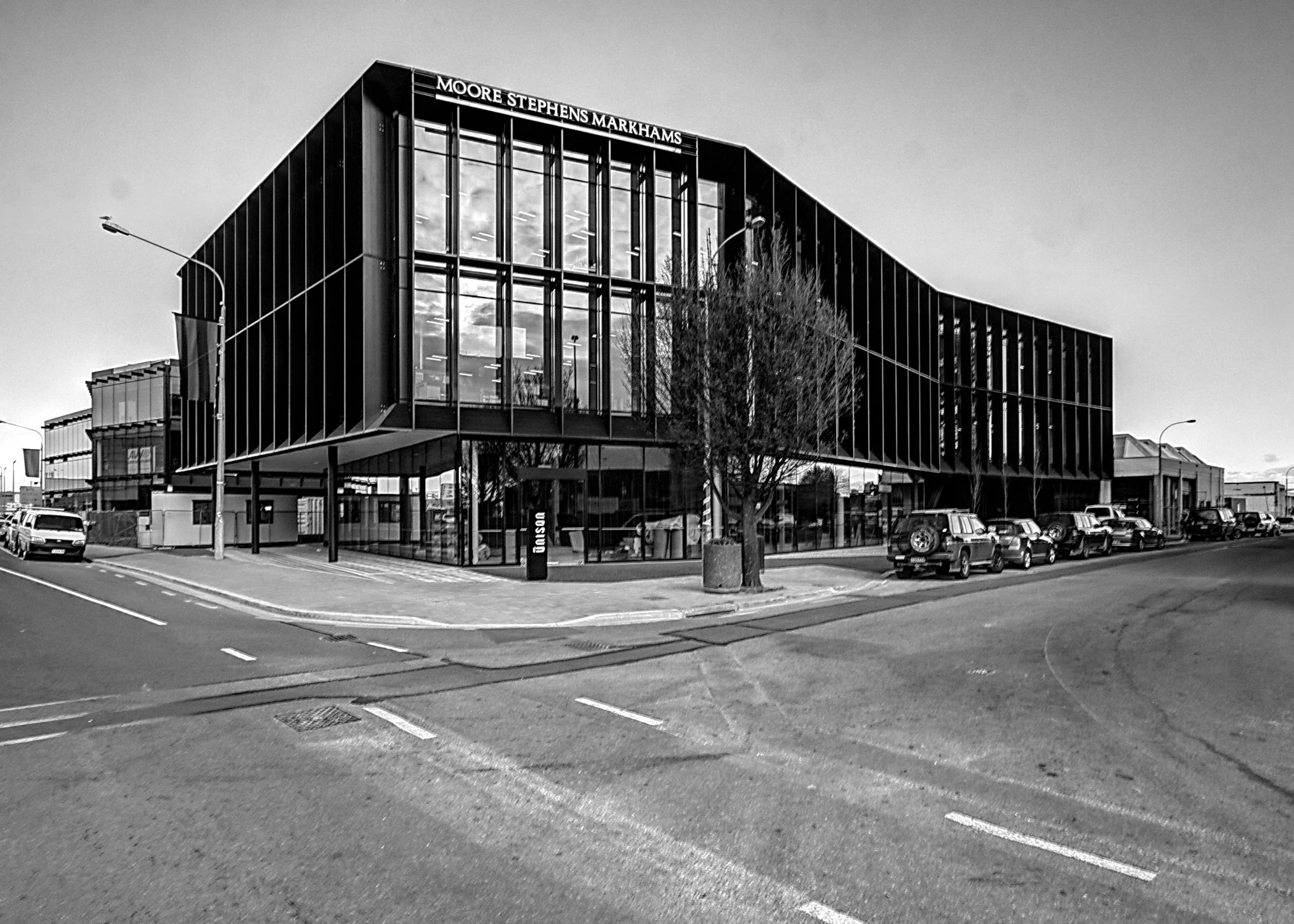 Black and white photo of a modern glass building with the sign 'Moore Stephens Markhams' on top, situated along a street with parked cars and a few leafless trees.