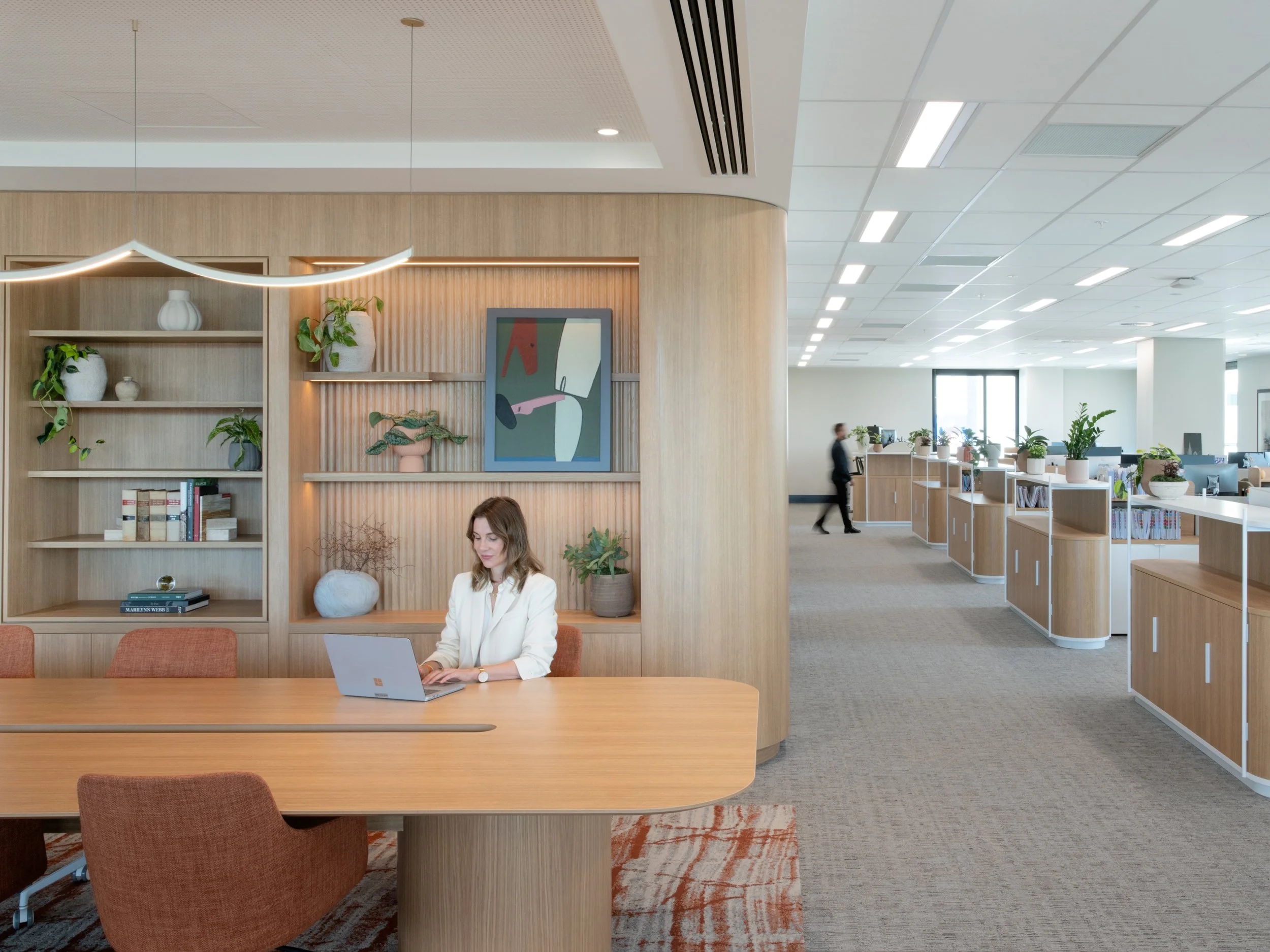 Modern office space with a woman working on a laptop at a wooden conference table, surrounded by bookshelves, potted plants, and artwork; open workspace with multiple desks and employees in the background.