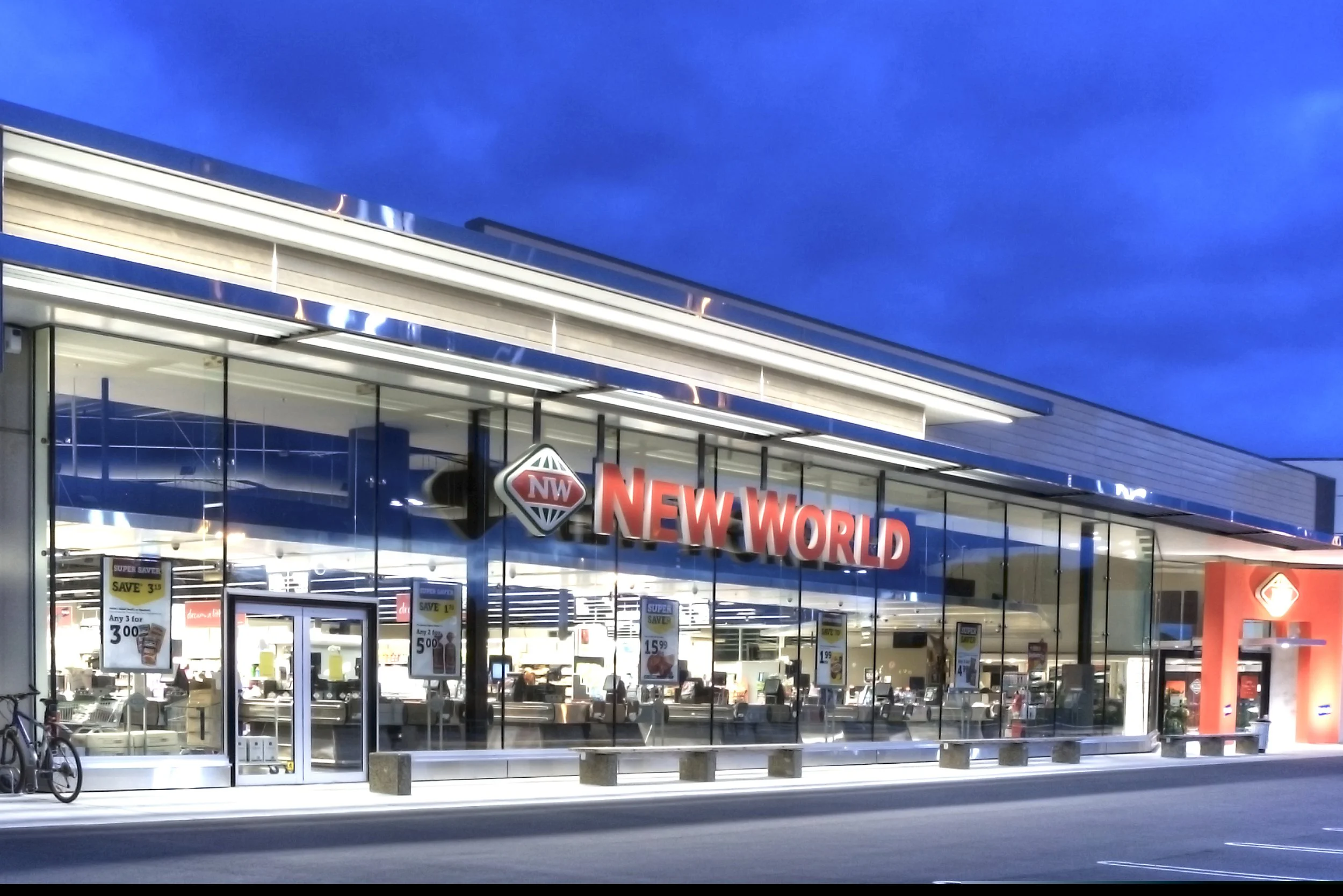 Exterior of a retail store named 'New World' with glass windows and blue signage, seen at dusk with a clear sky.