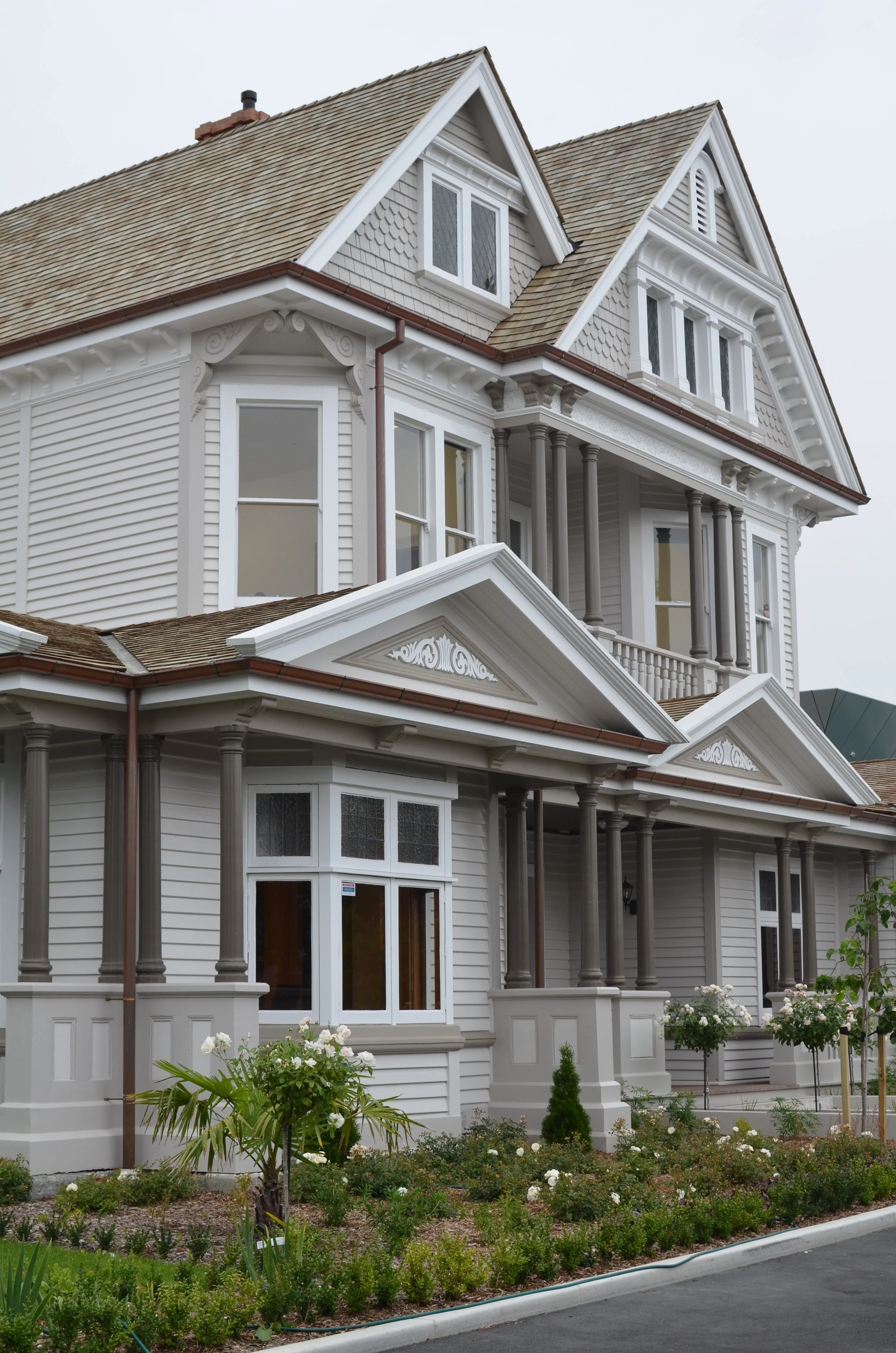 A large multi-story house with Victorian architectural details, painted white, featuring bay windows, decorative trim, and columns, with a well-maintained garden with flowers and bushes in front.