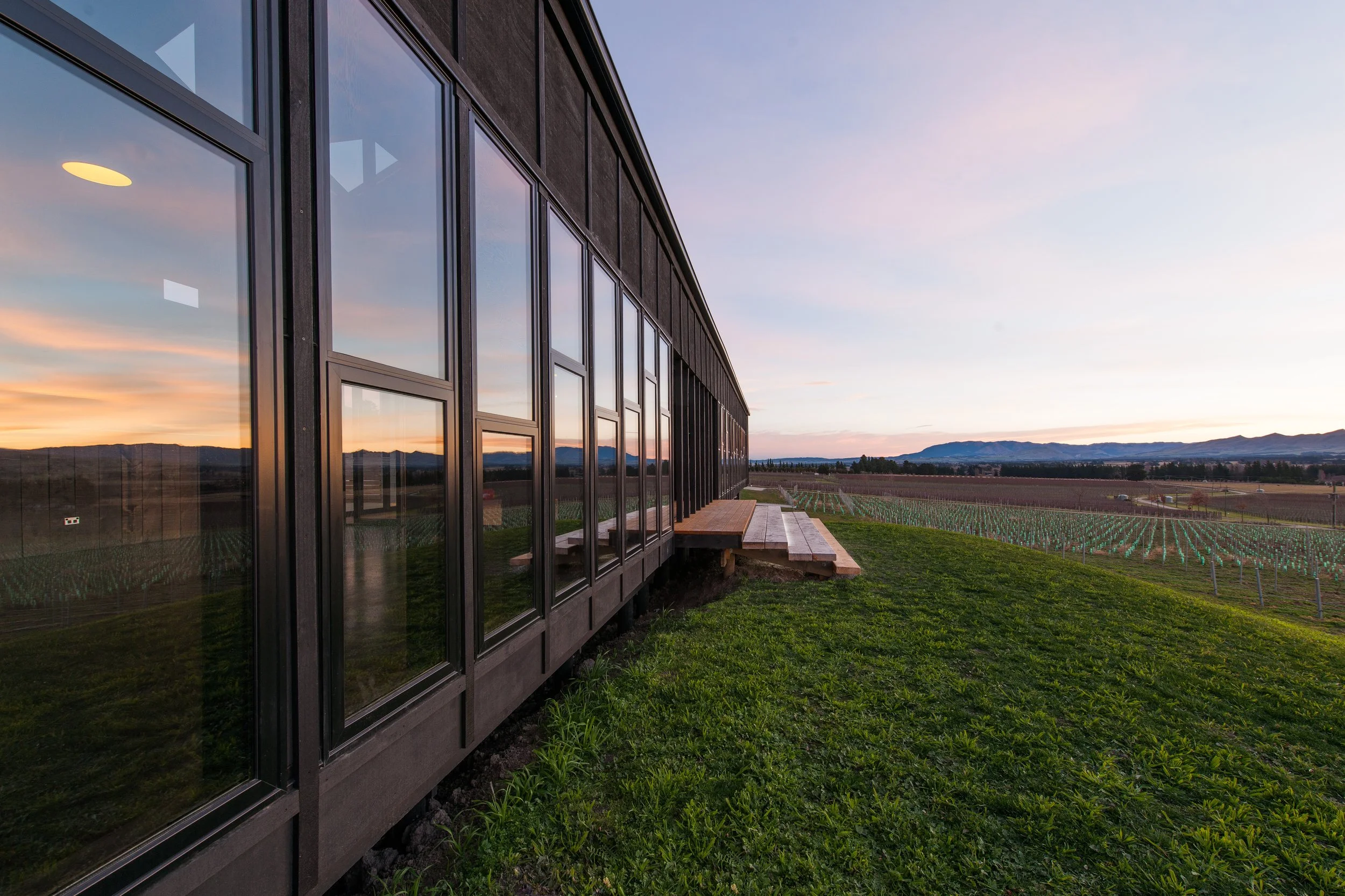 Sunset over a modern building with large glass windows, overlooking a landscape of fields and distant mountains.