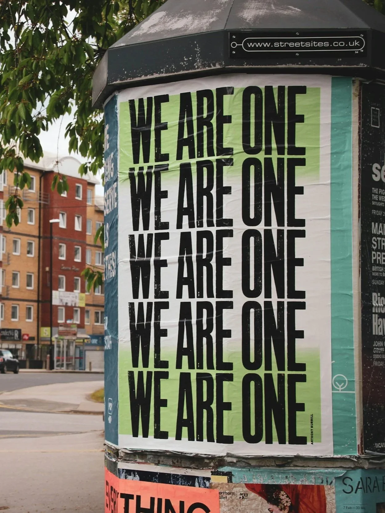 A poster with the repeated phrase "WE ARE ONE" in black text on a green and white background, attached to a street advertisement pillar in an urban setting with buildings and a tree visible.