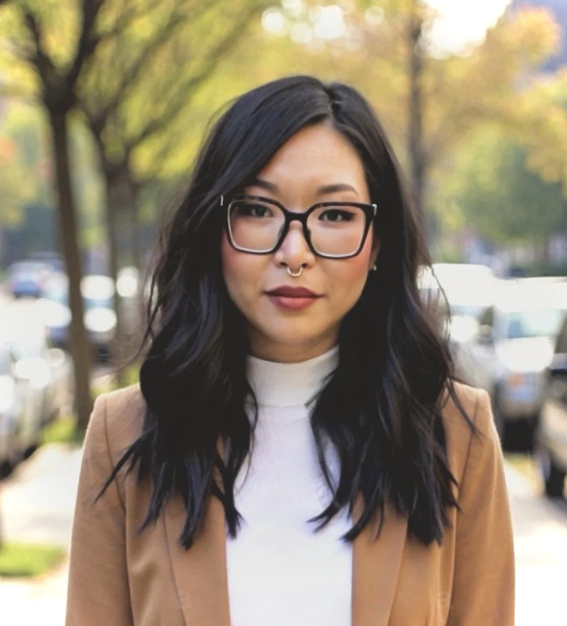 Portrait of a woman with dark wavy hair, wearing glasses, a septum piercing, a tan blazer, and a white turtleneck, standing outdoors with trees and parked cars in the background.