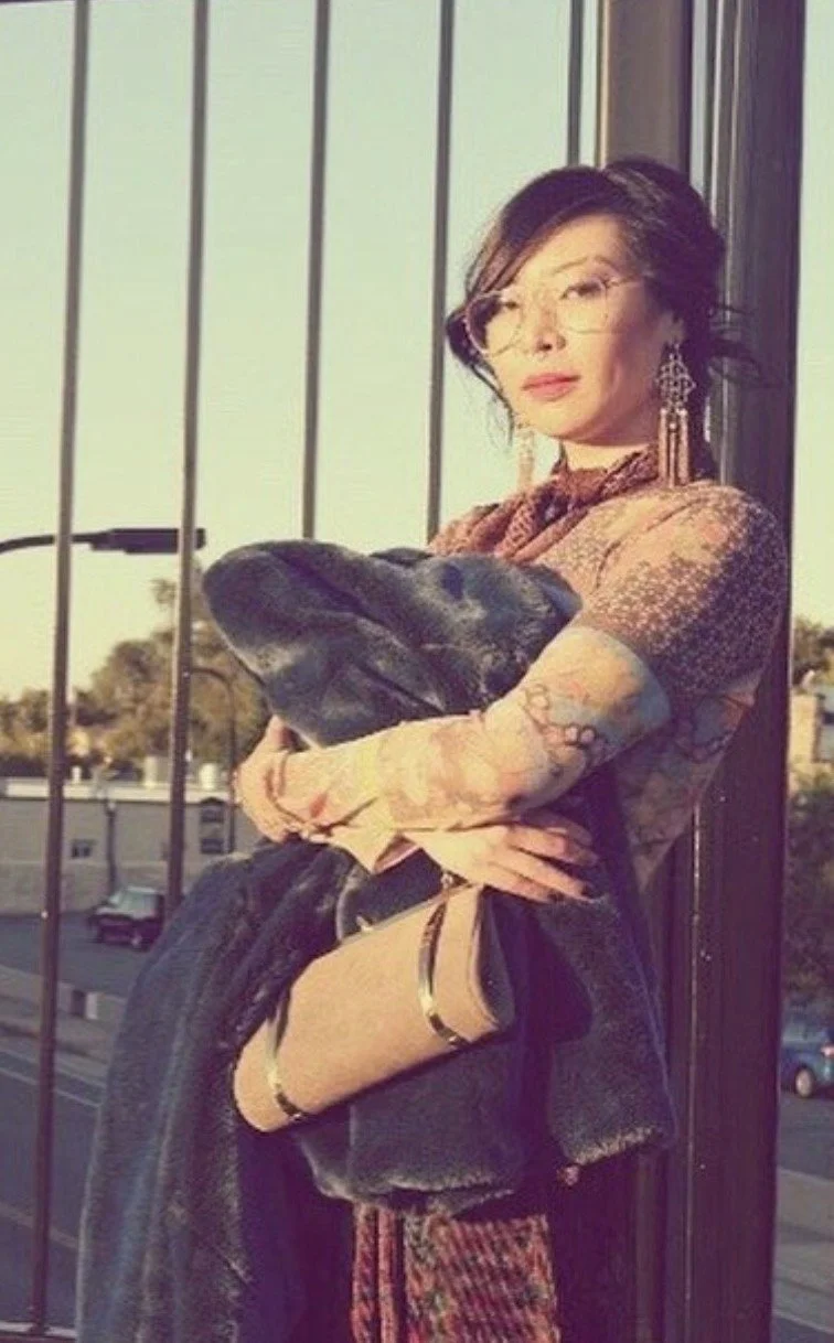 A woman with short dark hair, wearing glasses and earrings, is holding a large black dog in her arms. She is standing outdoors next to a metal railing, with a cityscape in the background.