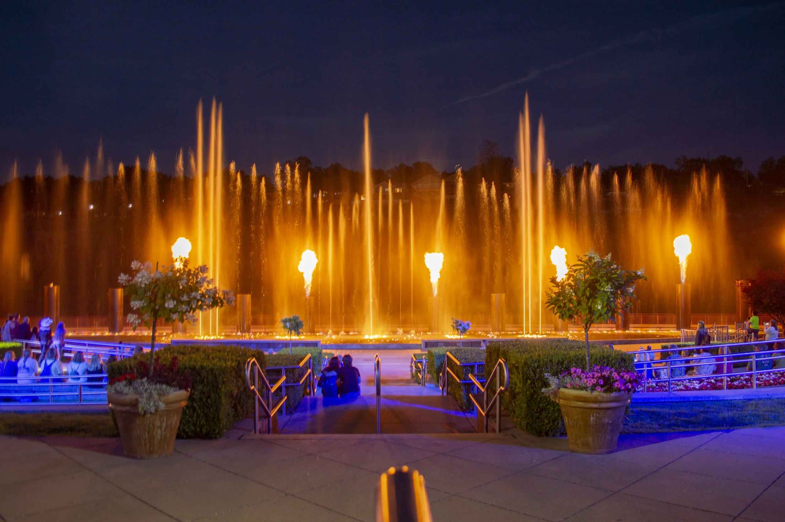A nighttime scene of a large illuminated fountain with orange water jets, surrounded by people sitting and walking, with potted plants and railings in the foreground.