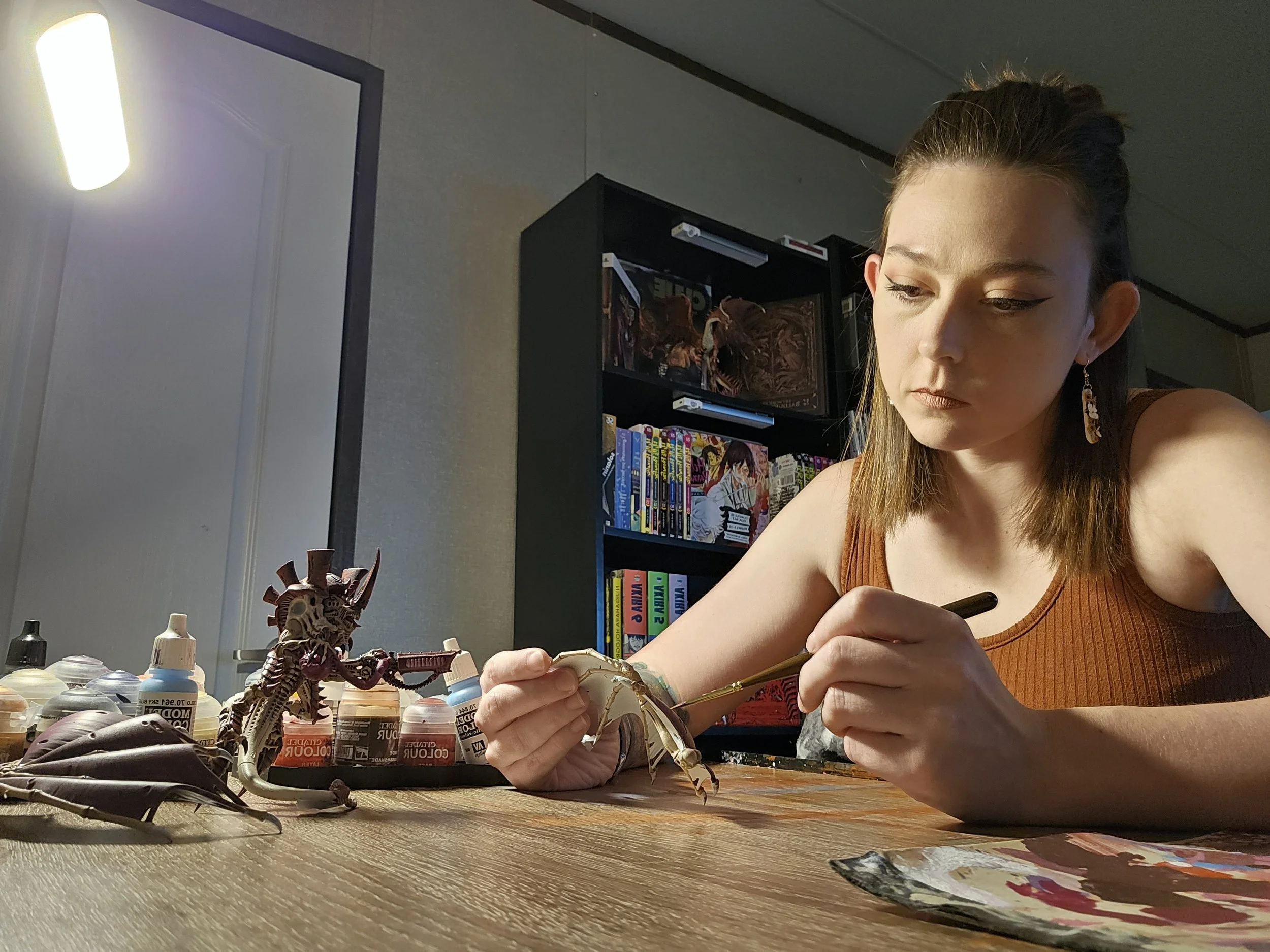 A woman working on assembling a model or figurine at a table with paint bottles and tools. She has long brown hair and is wearing a brown sleeveless top. Behind her, shelves with books and magazines are visible, along with a large TV or monitor.
