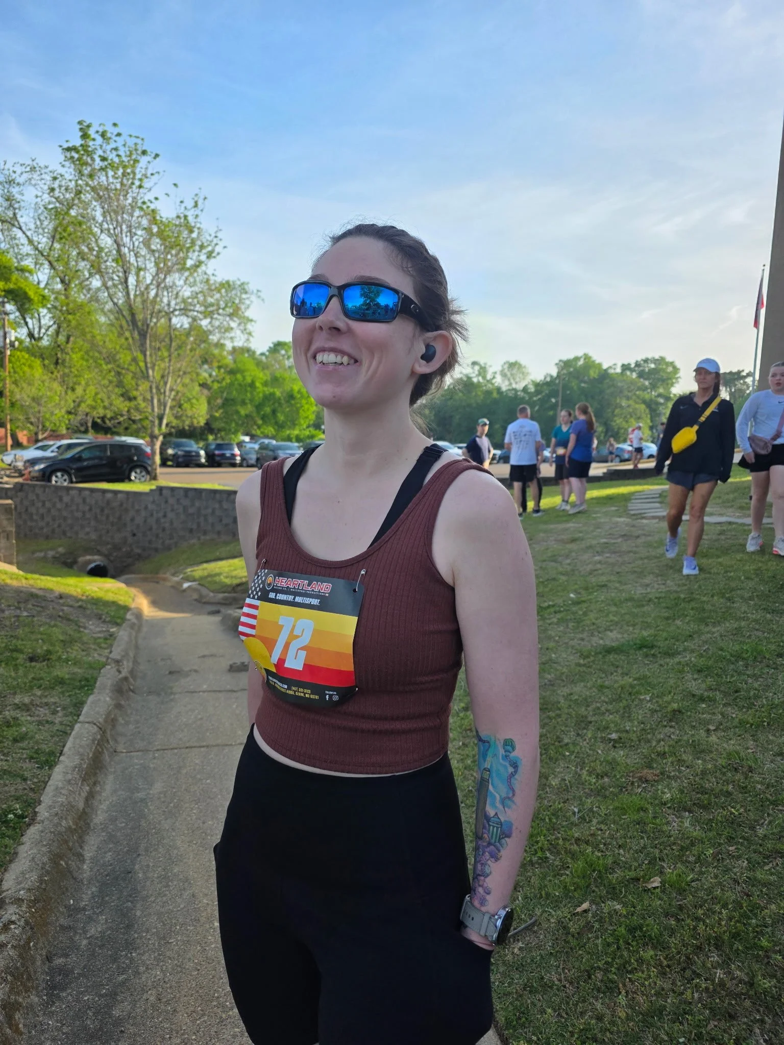 A female runner standing outdoors with a smile, wearing sunglasses, a marathon bib with the number 12, and black athletic clothing, with other runners and green trees in the background.