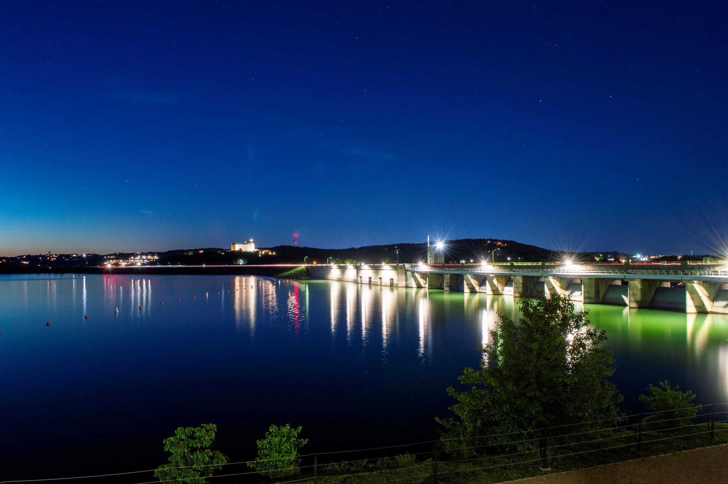 Nighttime view of a bridge over a body of water with city lights on the hills in the background. The sky is clear with visible stars.