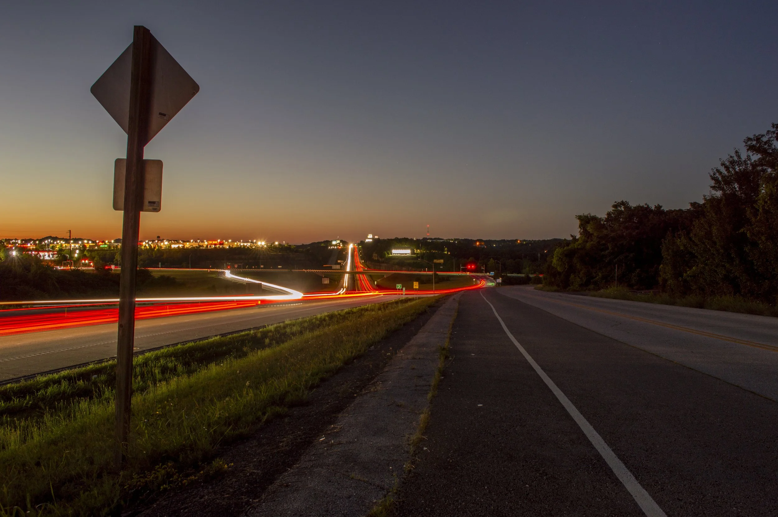 A highway at dusk with fading sunlight on the horizon, light trails from moving vehicles, a signpost on the left, trees on the right, and city lights in the distance.