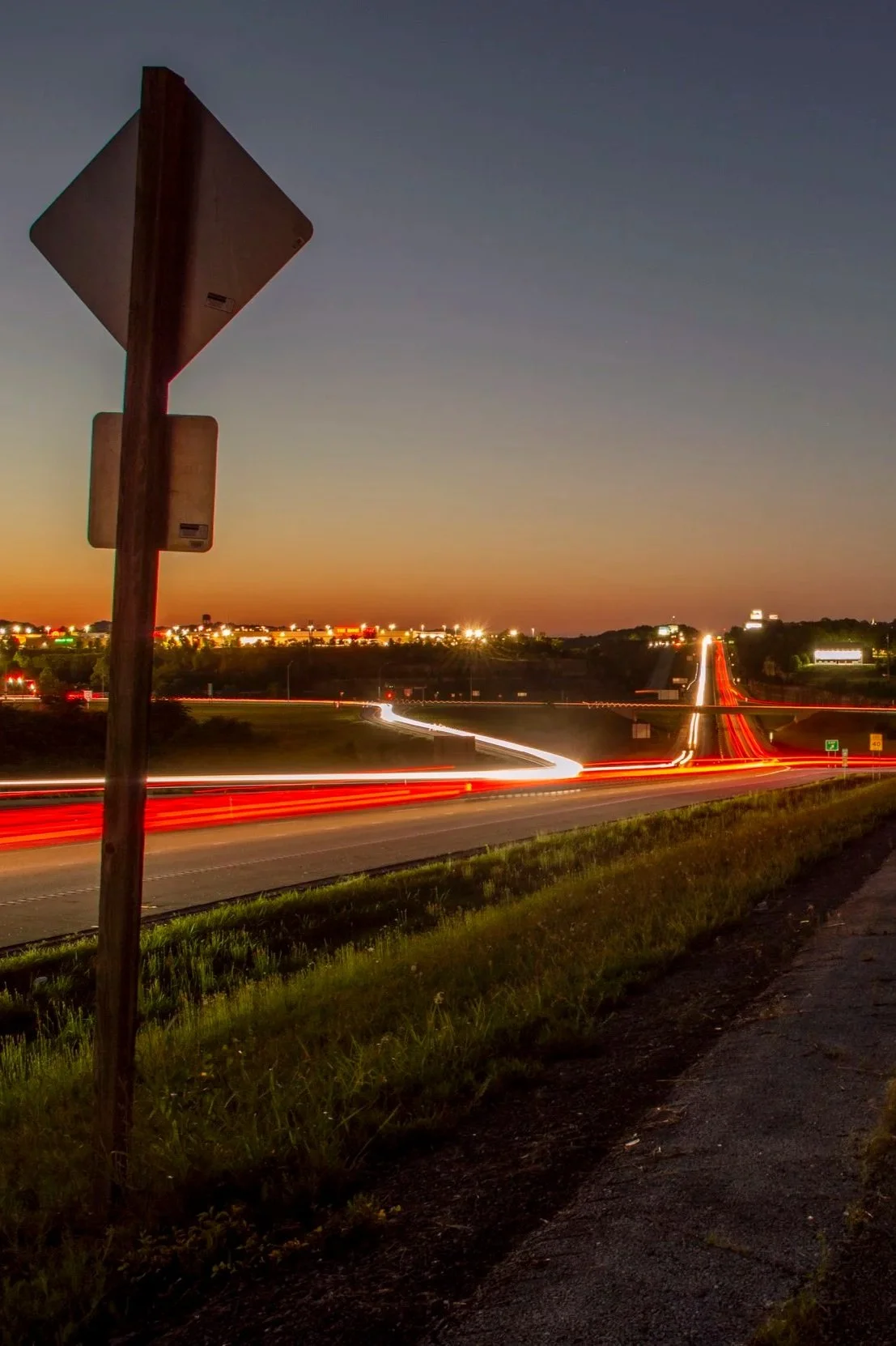 Long exposure photograph of a highway at dusk, showing streaks of red and white lights from moving vehicles, with a signpost in the foreground and a cityscape with illuminated buildings in the background.