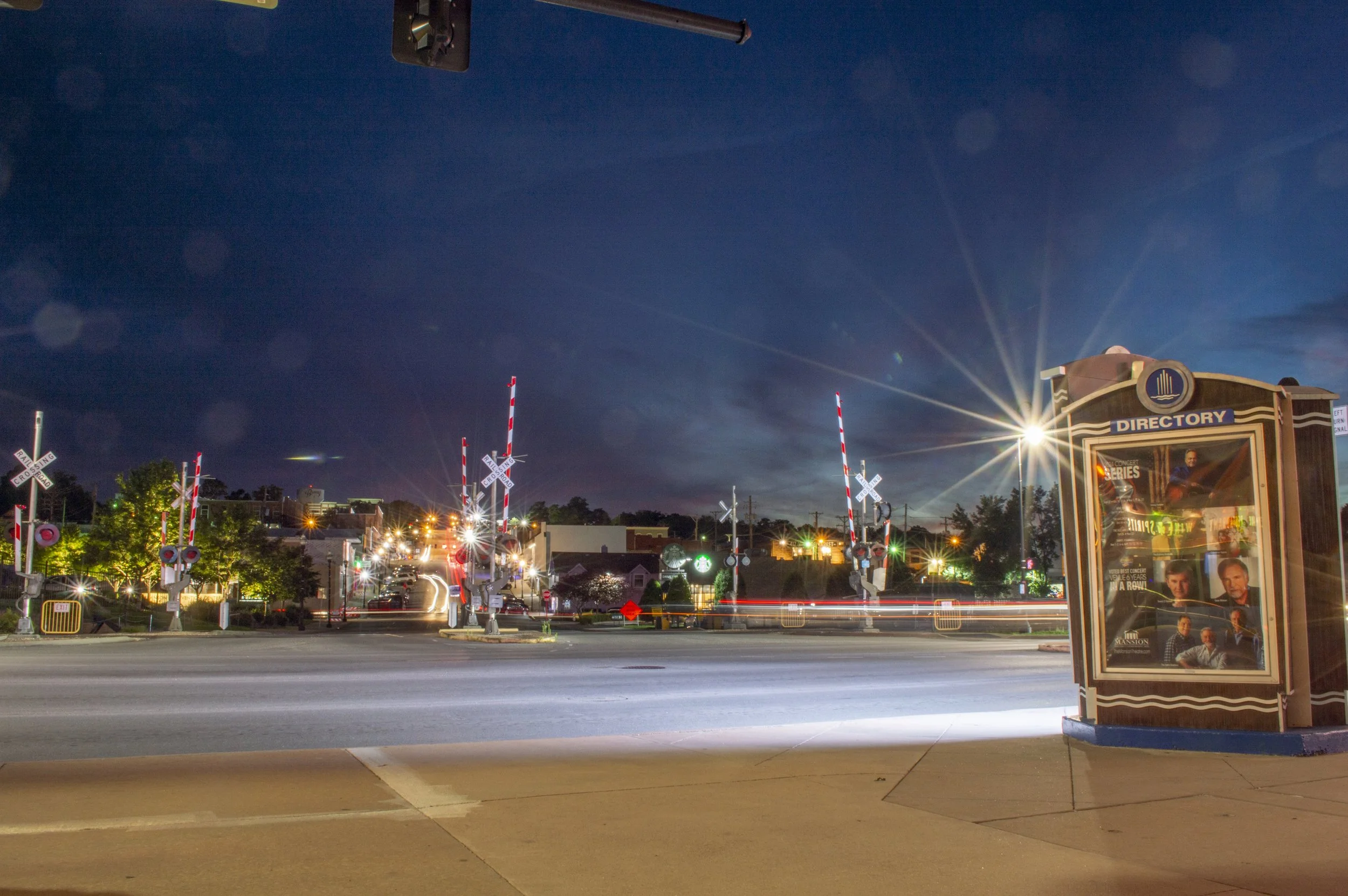 A nighttime street scene with bright city lights, a directory kiosk on the right, railroad crossing signs, and a clear dark blue sky with some clouds.