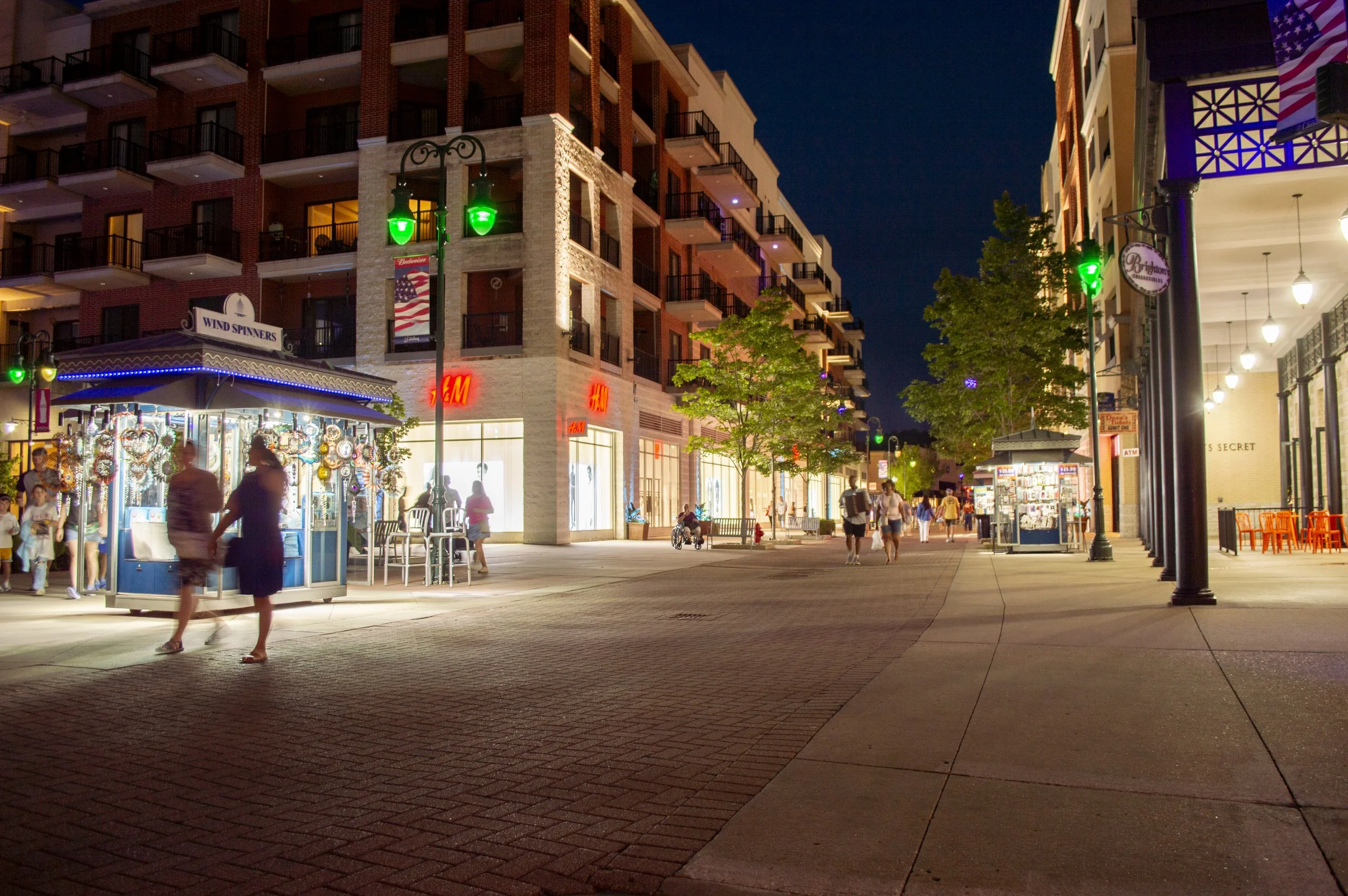Nighttime street scene with illuminated shops, including a jewelry stand and a clothing store, trees, and people walking, some with shopping bags, under streetlights. Buildings with balconies line the street.