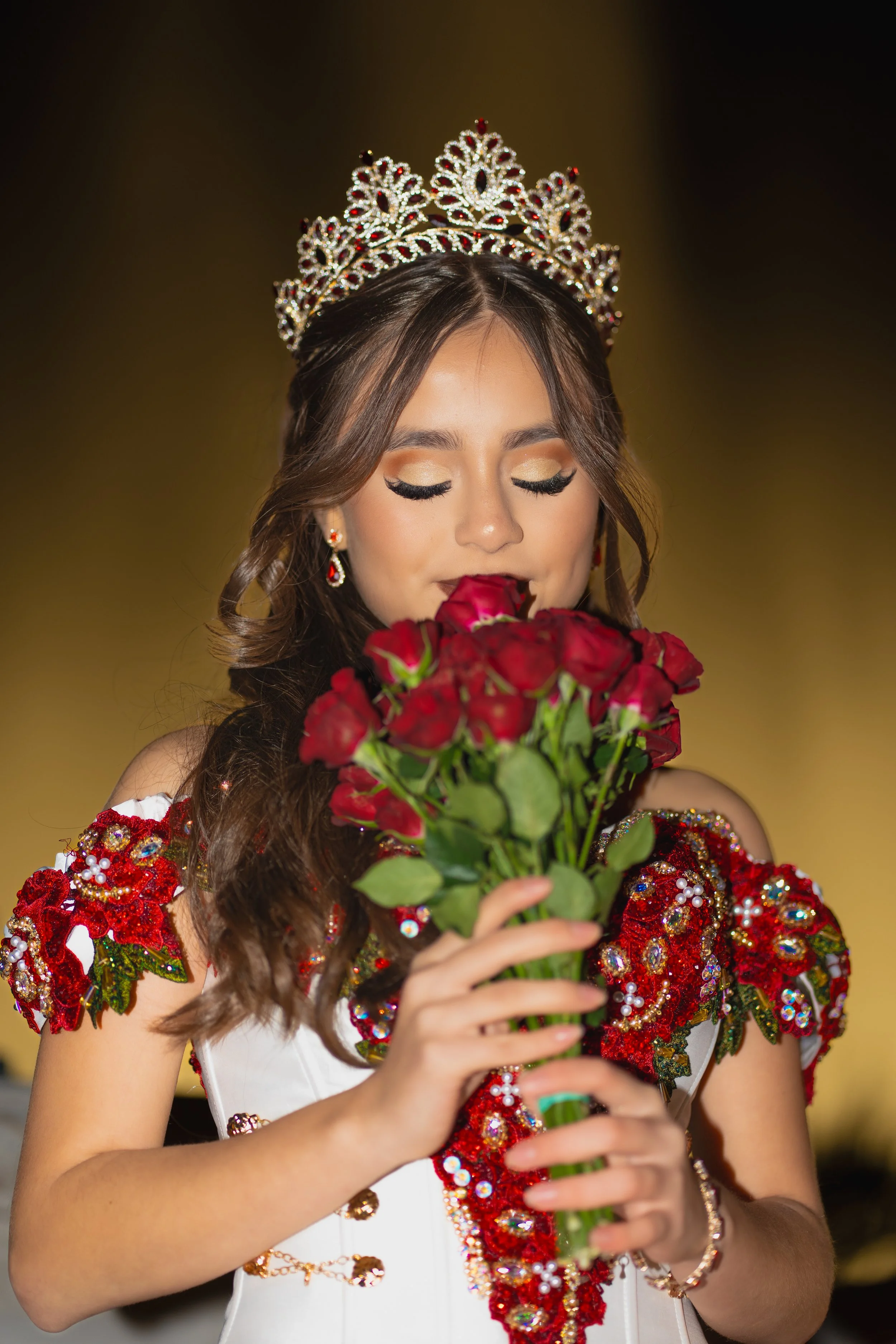 A woman wearing a sparkling tiara and a colorful, ornate dress with red, gold, and green embellishments holds a bouquet of red roses with her eyes closed.