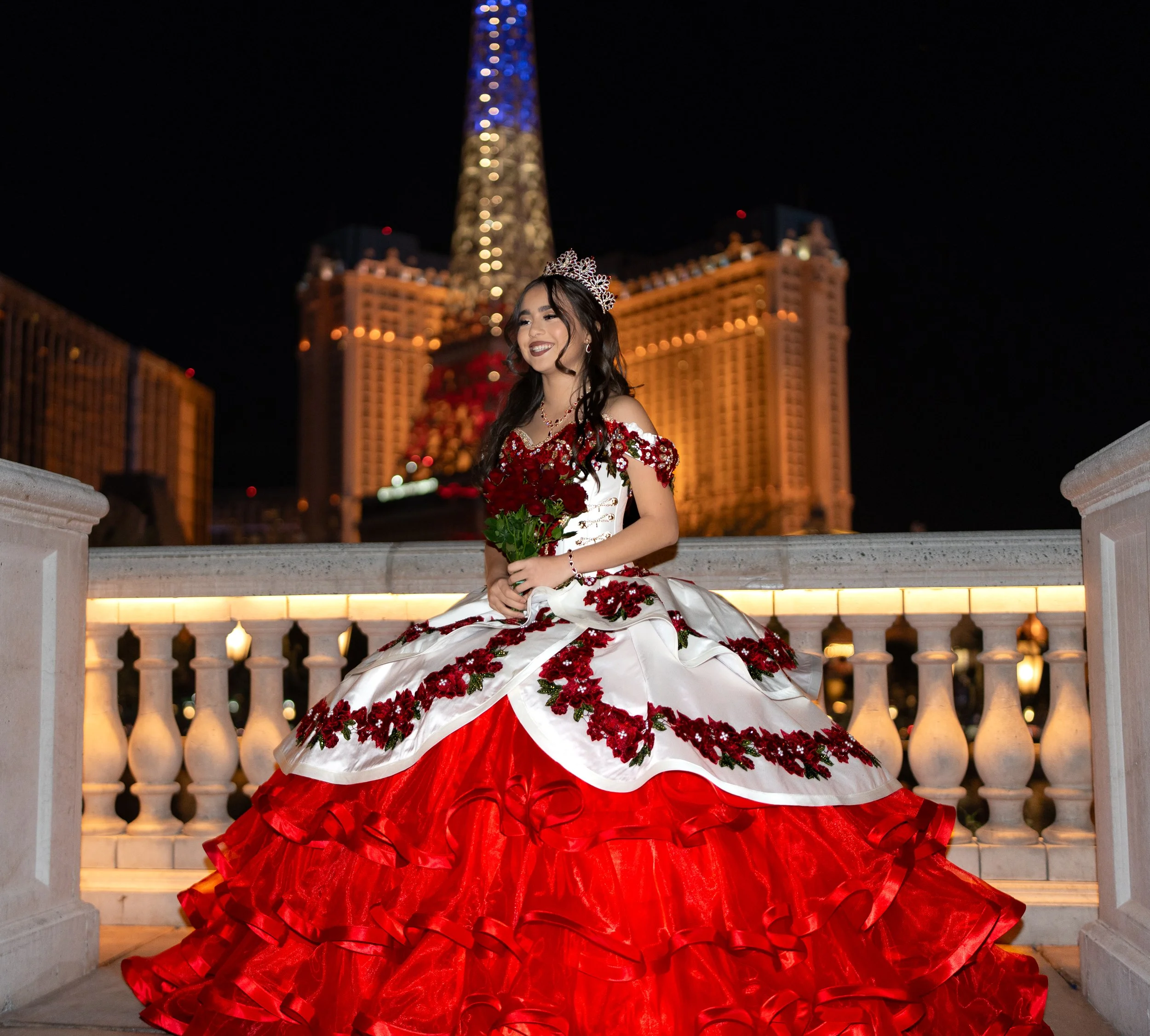 Young woman wearing a sparkling tiara and a floor-length red gown with floral embroidery, sitting on a balcony railing at night with a lit hotel or casino building and a tall tower in the background.
