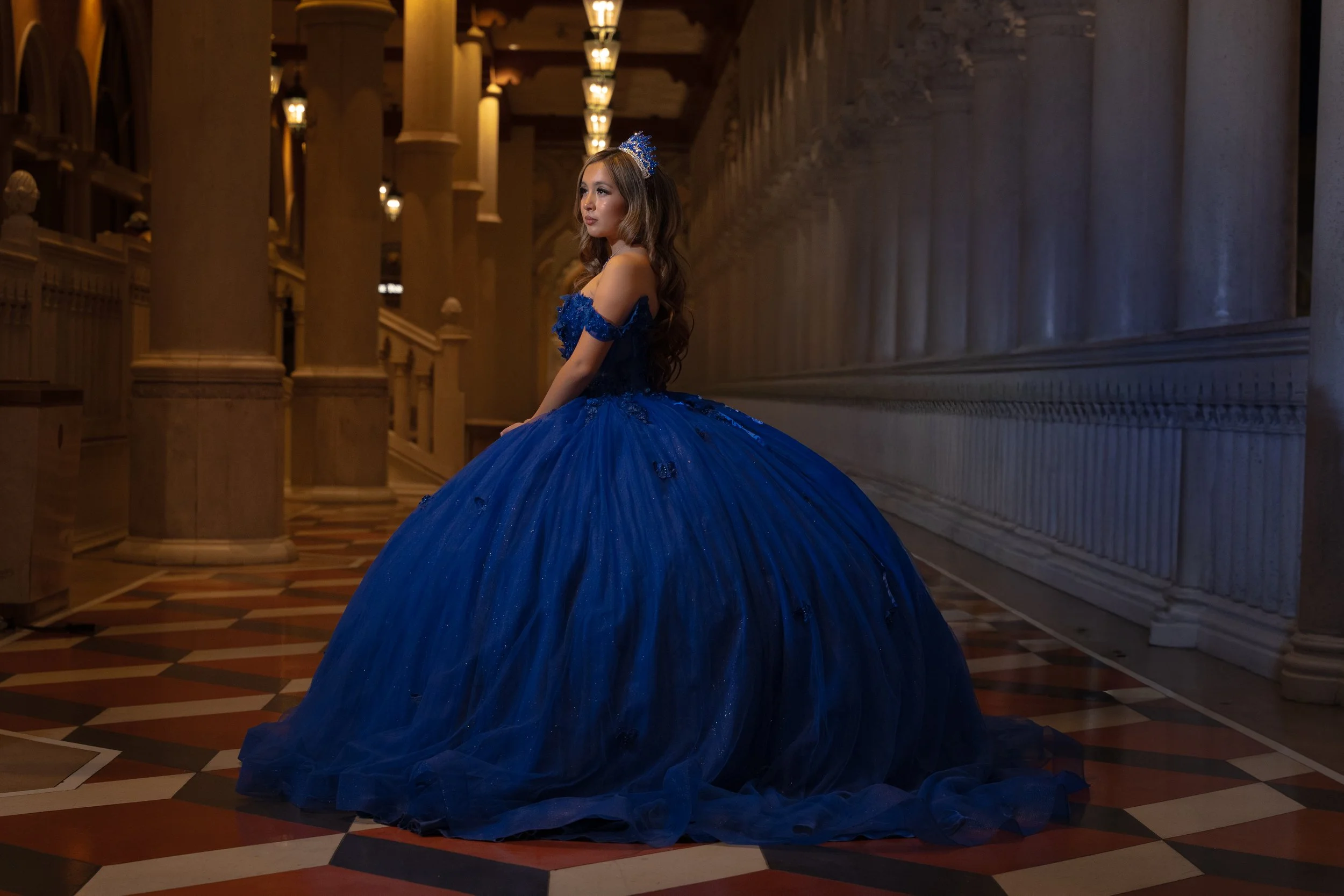 A young woman in a royal blue ball gown with off-shoulder ruffles and sequin embellishments, wearing a matching headpiece, standing in an elegant, warmly lit hall with tall columns and ornate lighting fixtures.