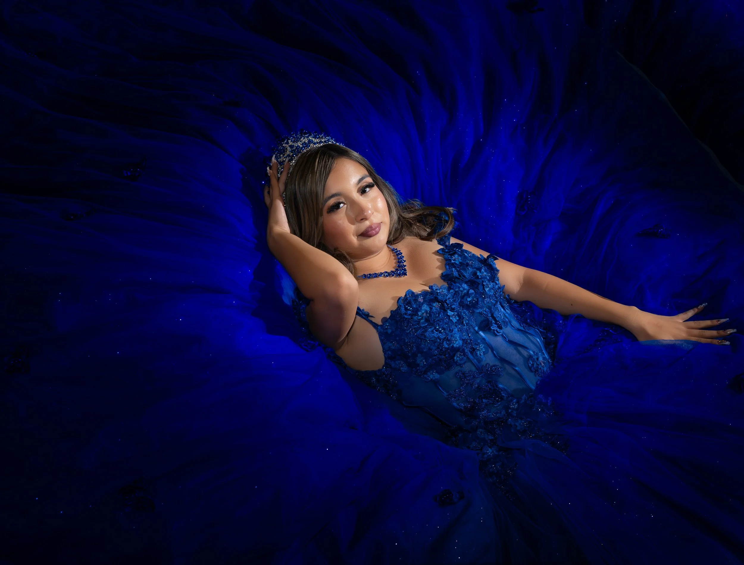 A woman lying on a dark blue velvet fabric with her head resting on her hand, wearing a blue lace dress and a silver tiara.