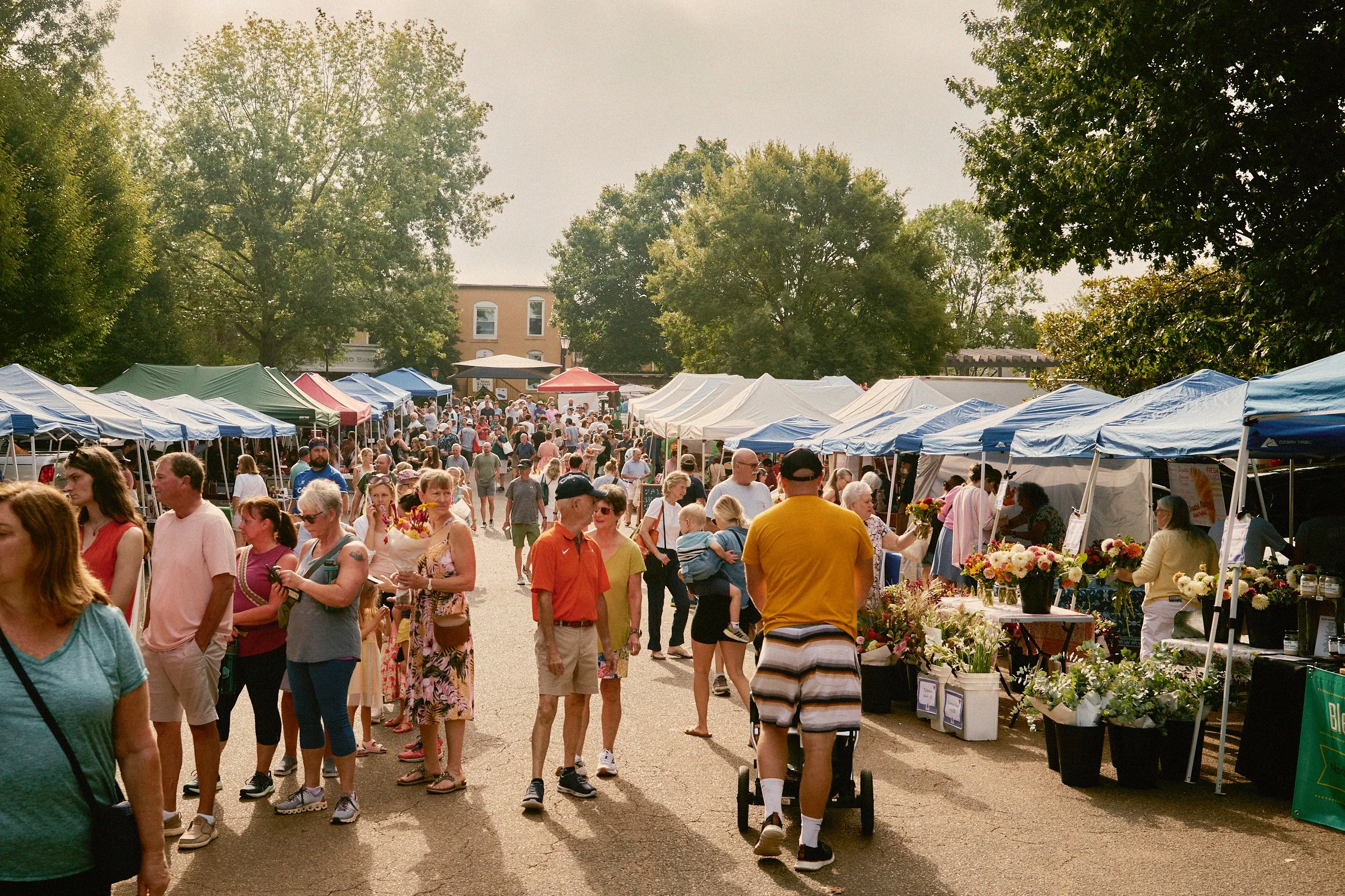 Outdoor farmers market with tents and a crowd of people shopping and walking among vendor stalls on a sunny day.