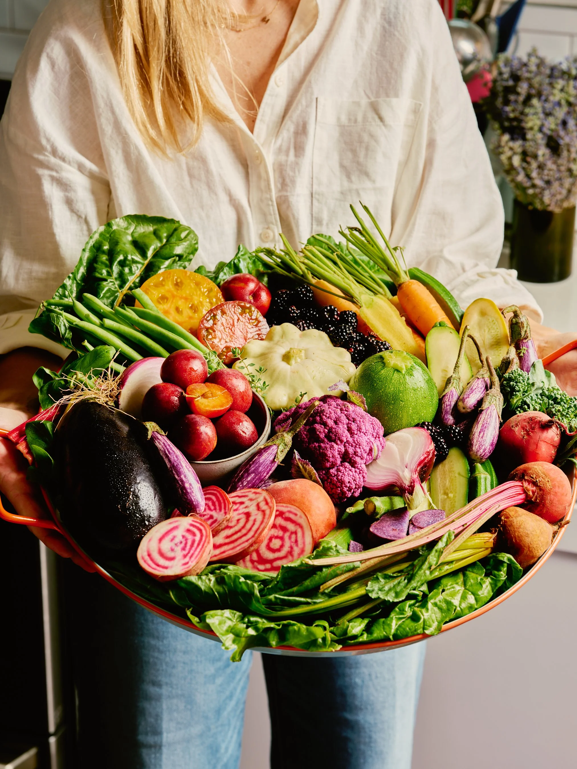 Person holding a large decorative basket filled with colorful fresh vegetables and fruits, including eggplant, radishes, cauliflower, carrots, beets, and greens.