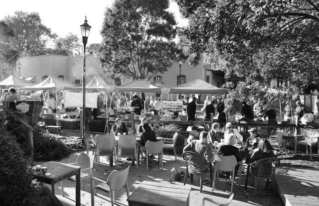 Outdoor market or fair with numerous tents, tables, and people browsing and sitting around. Trees and buildings are in the background, with a streetlamp visible.