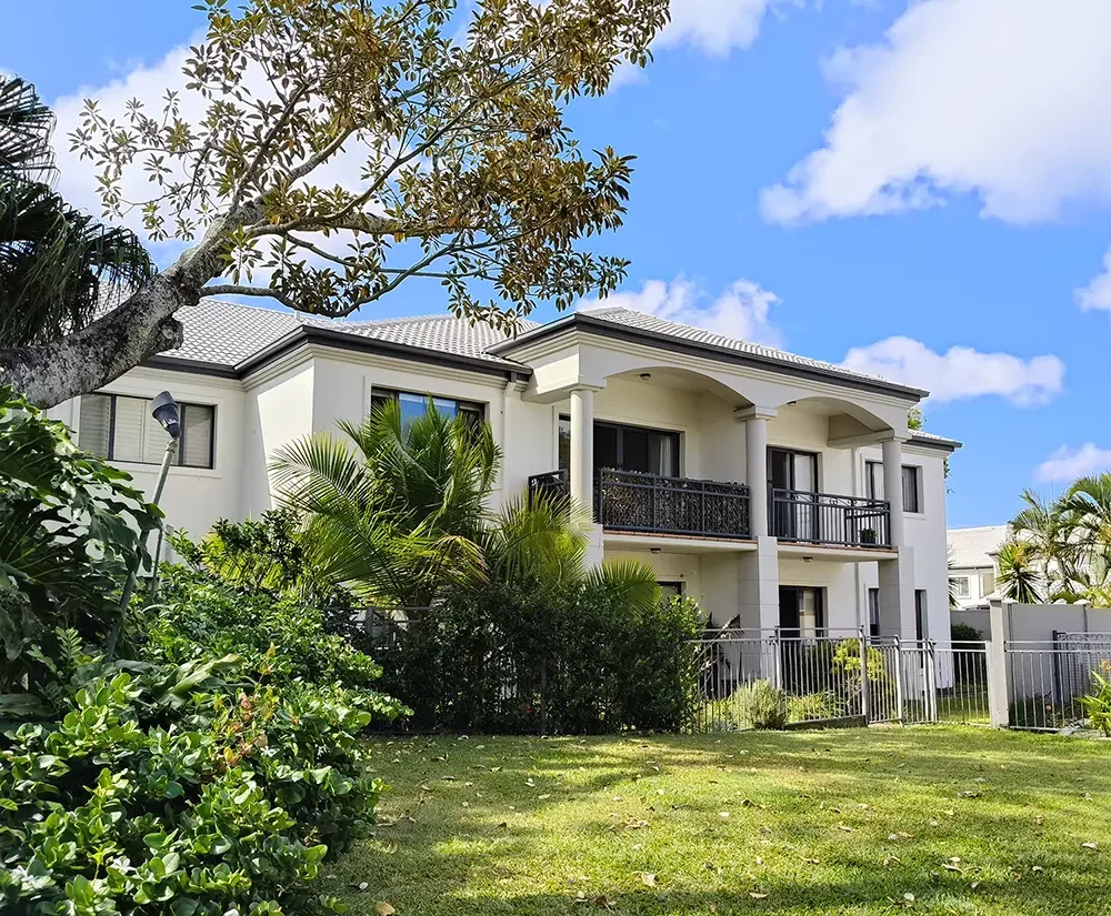A white with charcoal trim two-story townhouse in a Gold Coast body corporate estate, surrounded by green bushes and palm trees, under a blue sky with clouds.