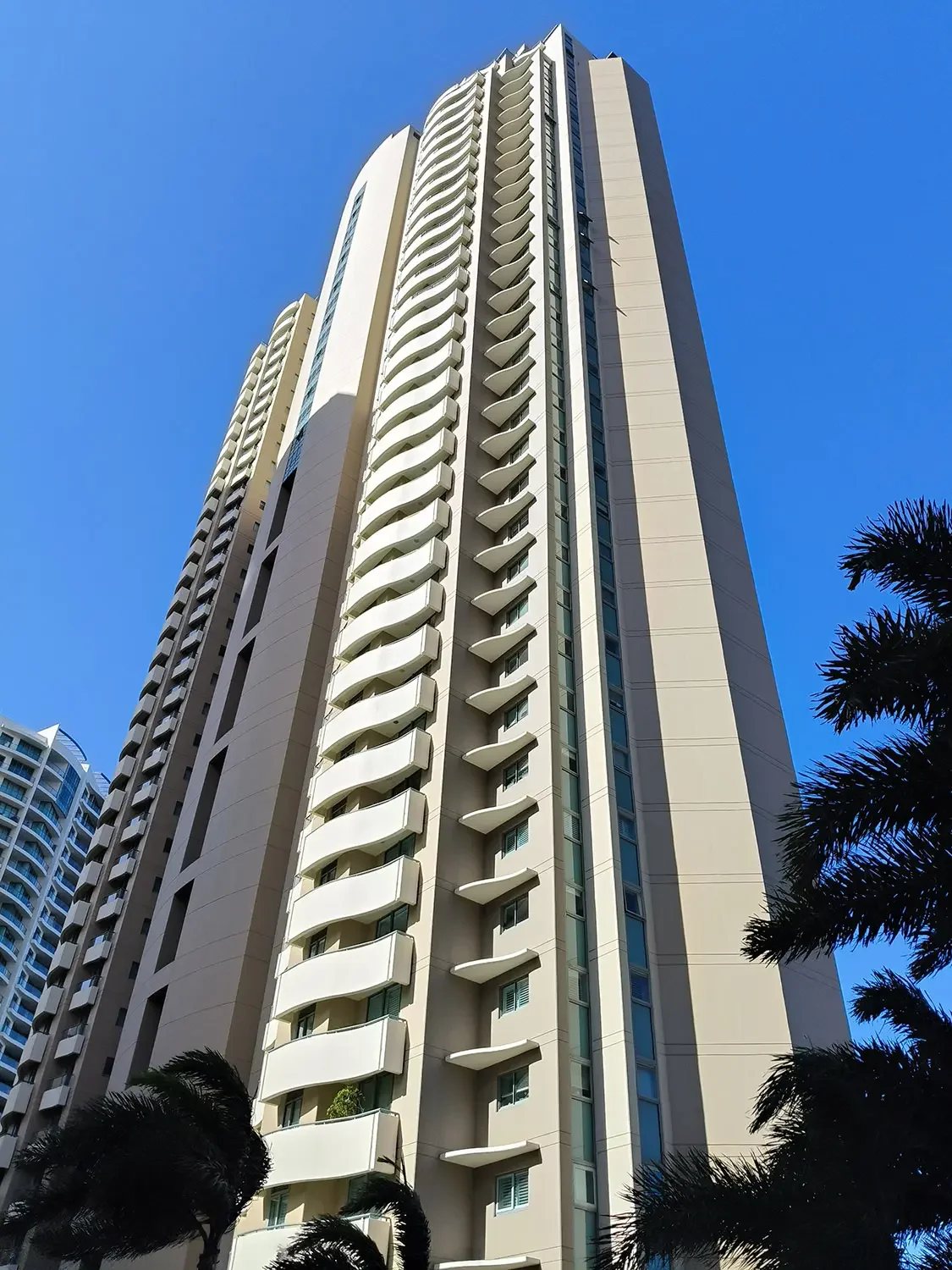 A tall modern high-rise building in South East Queensland with a refreshed colour scheme, against a clear blue sky, with palm trees in the foreground.